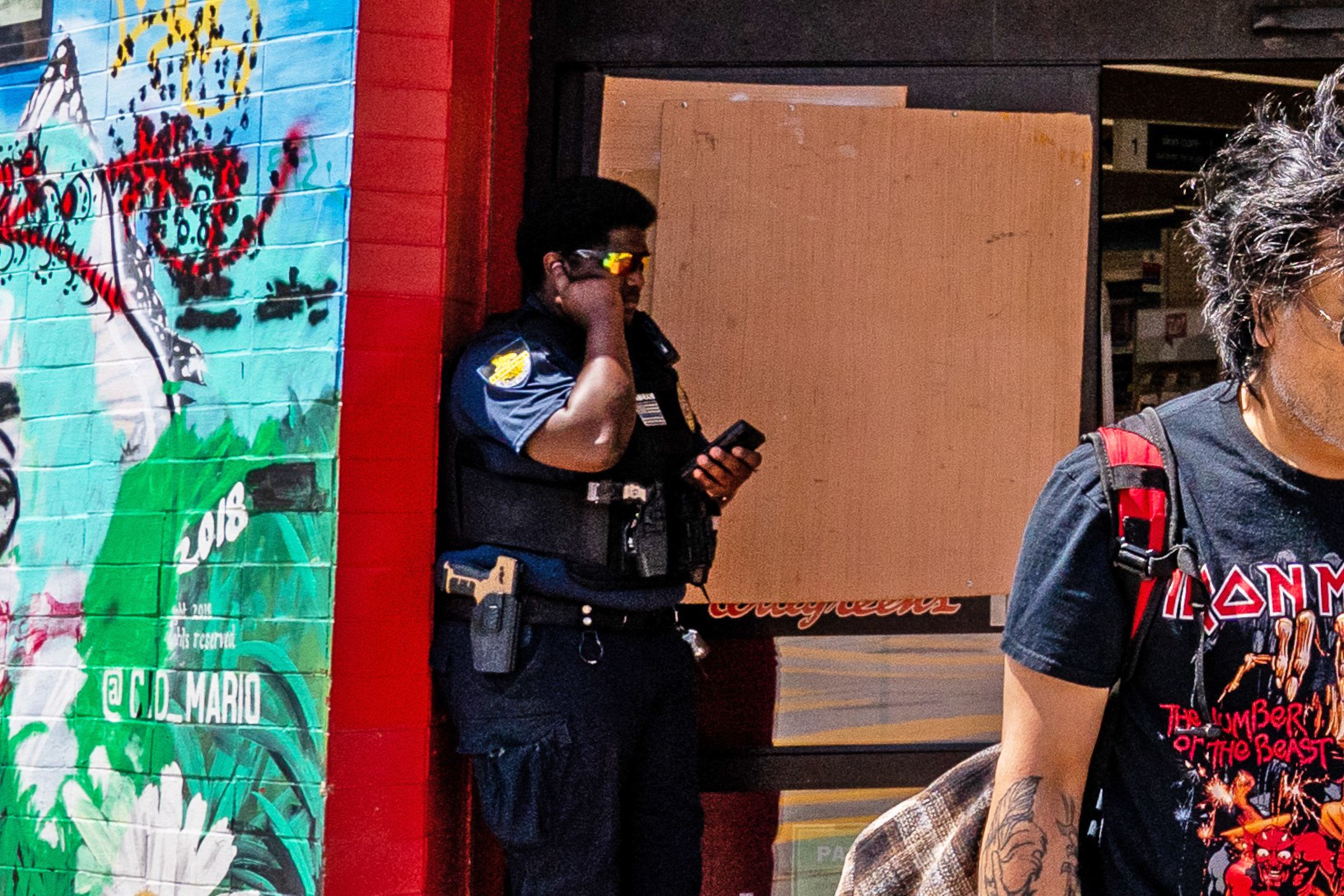 A police officer wearing sunglasses and a vest stands against a red wall with graffiti, talking on the phone, while a man with tattoos wearing an Iron Maiden shirt walks by.