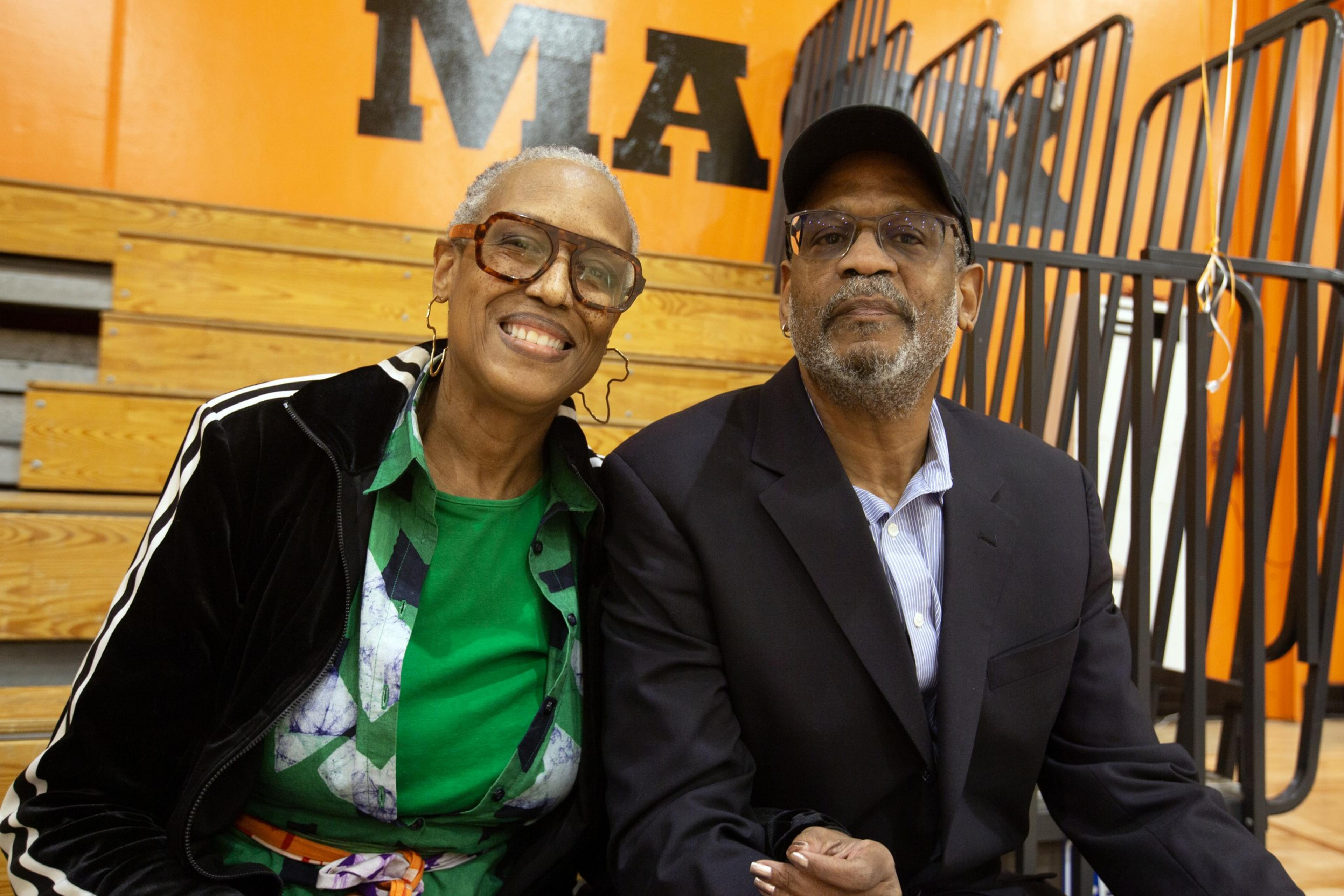 An elderly couple is sitting together, smiling. The woman wears glasses and a green shirt, while the man is in a suit and cap. They're indoors, on bleachers.