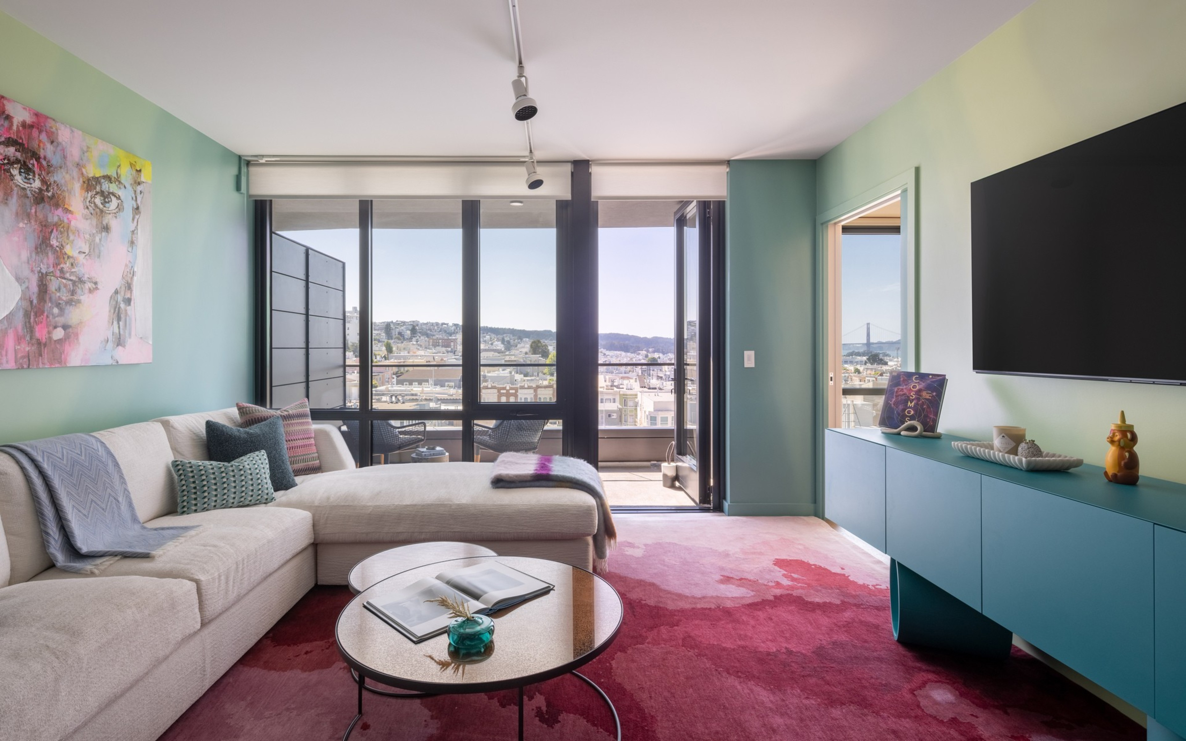 A modern living room with a beige sectional sofa, pink and red area rug, round glass coffee table, blue sideboard, wall-mounted TV, and large windows leading to a balcony.