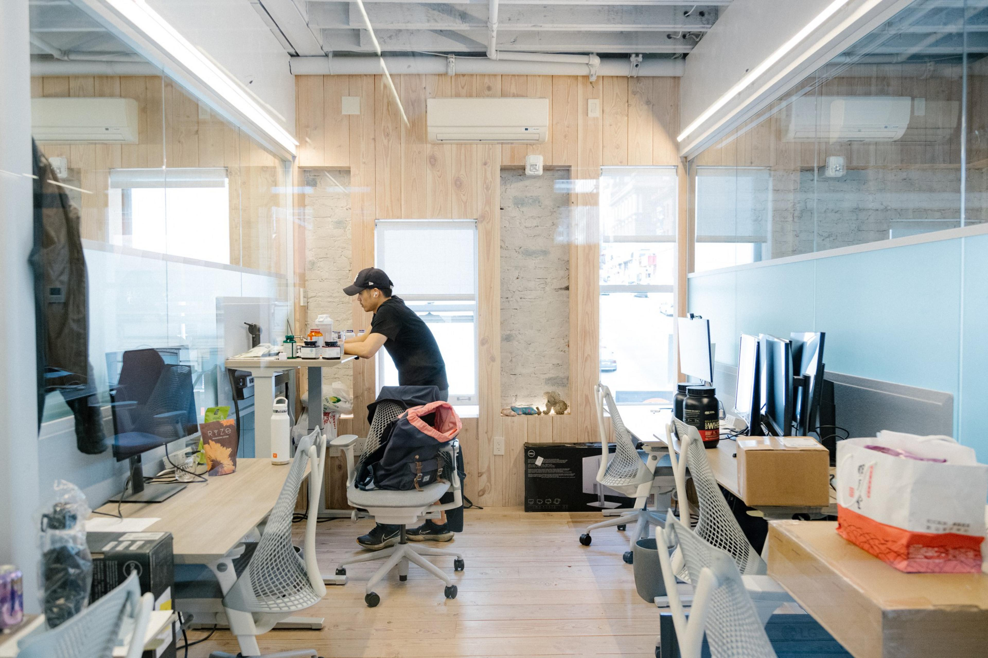 A man wearing a black cap and shirt leans over a standing desk in a bright office with wooden walls, multiple monitors, and ergonomic chairs.