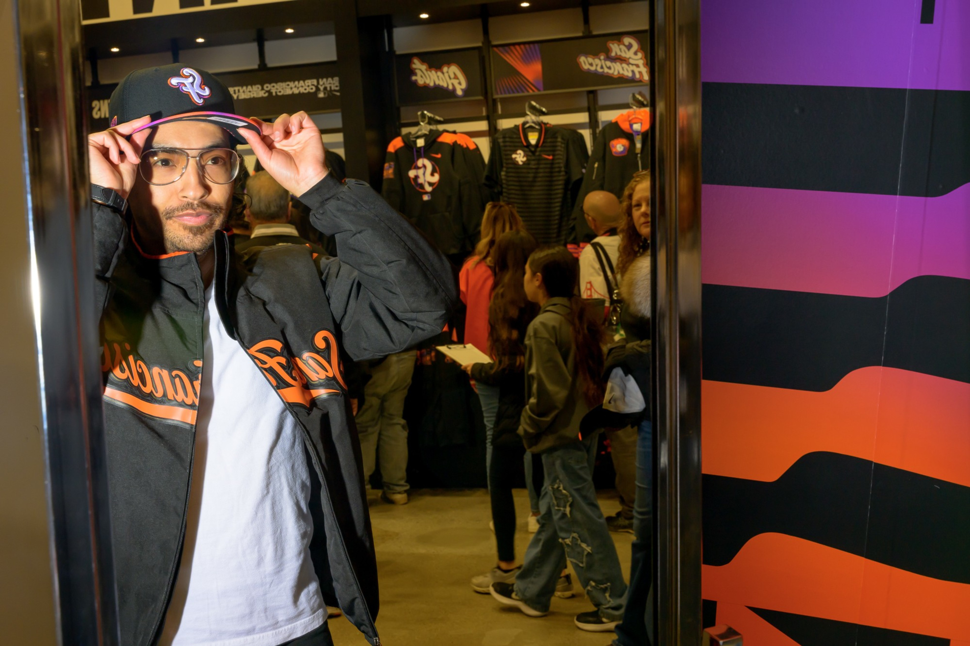 A person in a San Francisco Giants jacket and cap adjusts their hat in a store. Behind them, people browse baseball apparel. A colorful wall is partially visible.
