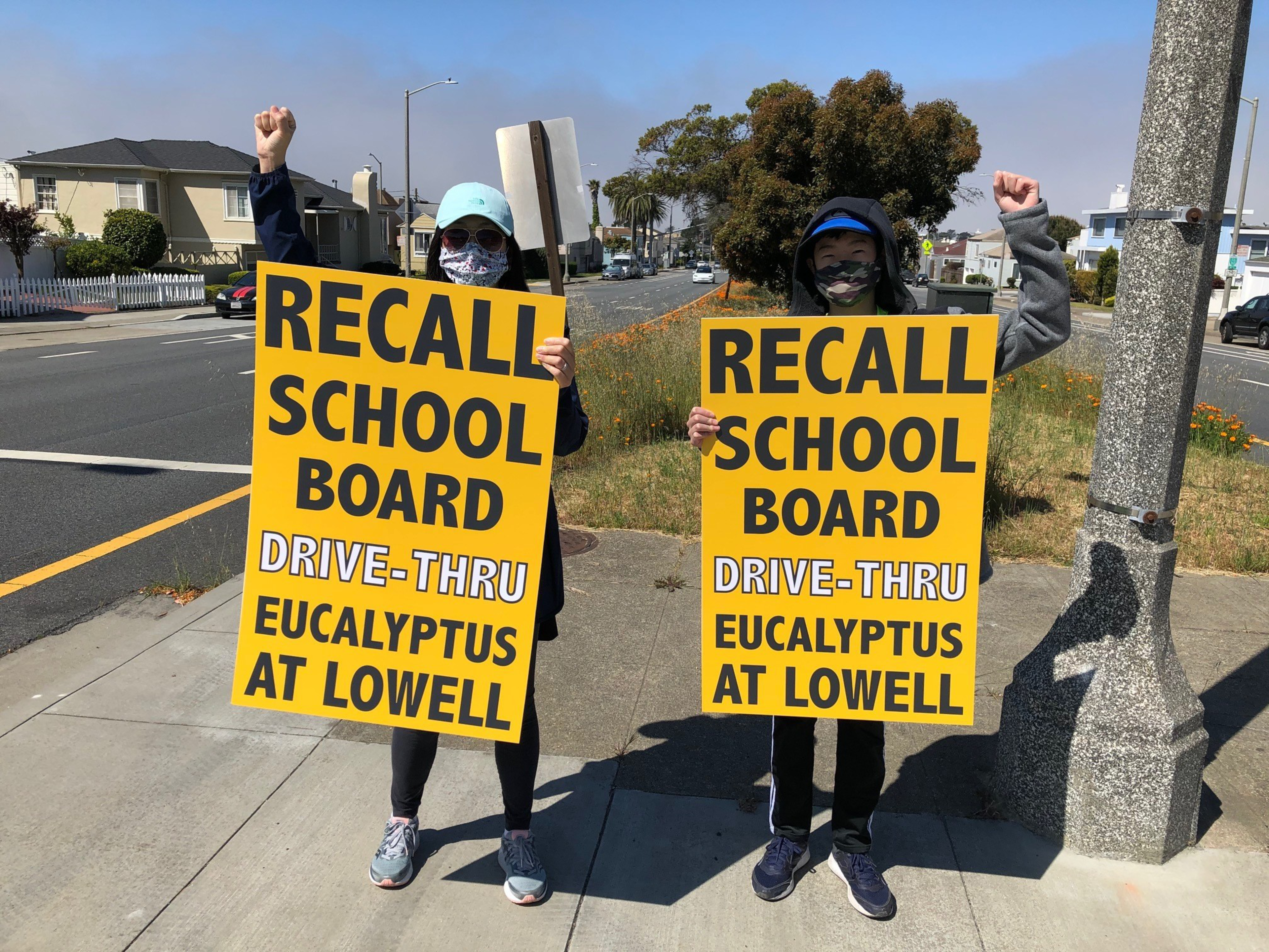 Two people wearing masks and hats hold yellow signs saying “RECALL SCHOOL BOARD DRIVE-THRU EUCALYPTUS AT LOWELL” while raising fists on a sidewalk.