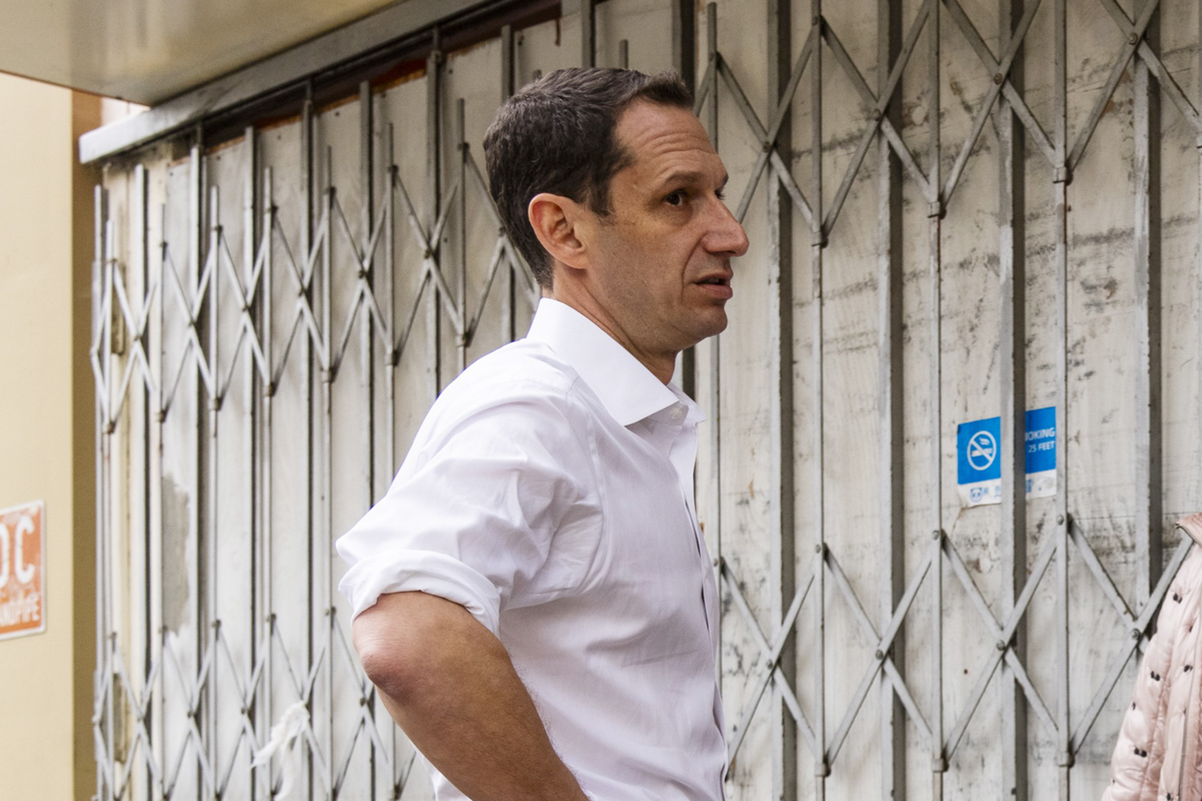 A man in a white shirt stands in front of a closed metal gate with his hands on his hips. A blue ""no smoking"" sign is visible in the background.
