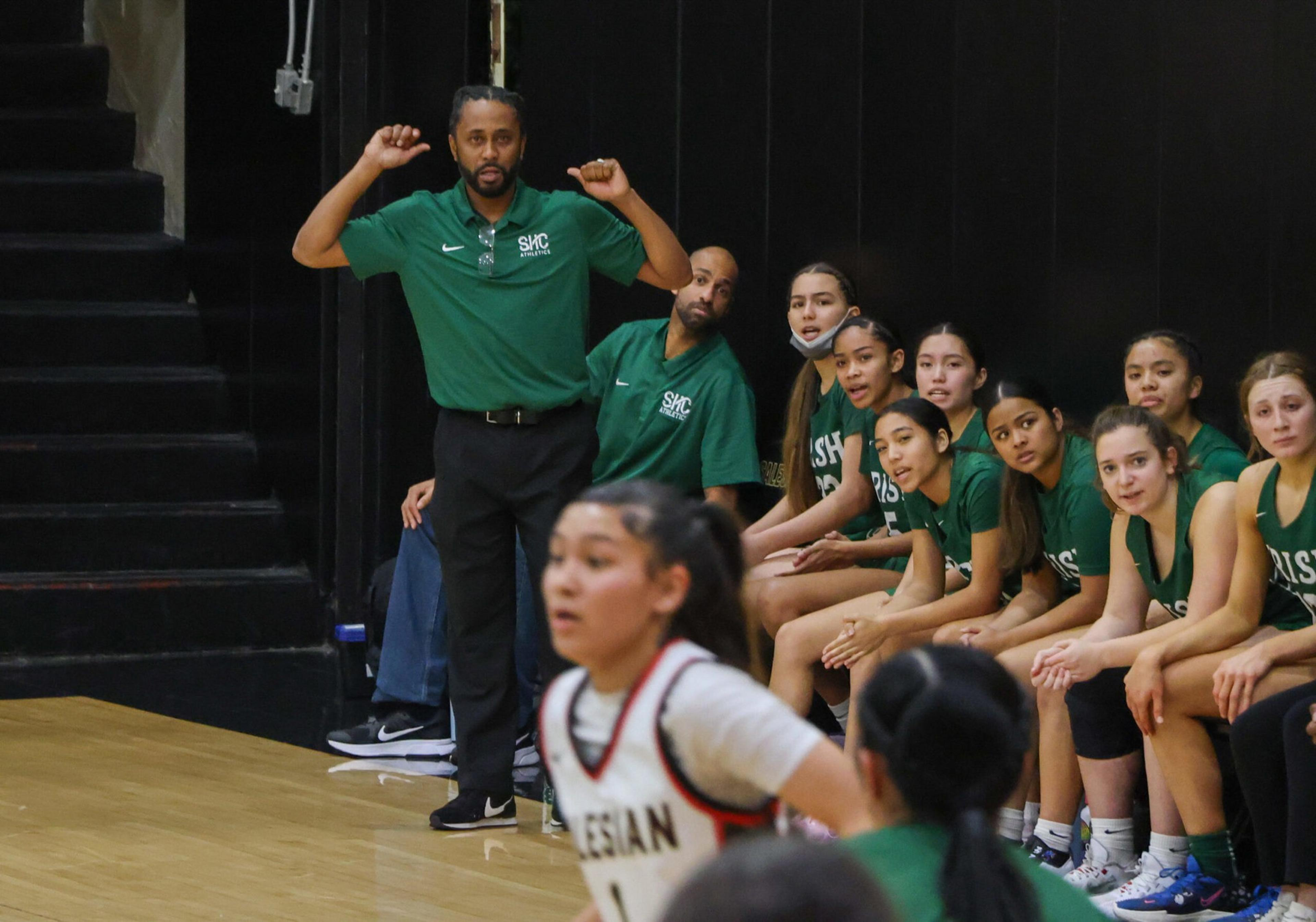 A basketball coach gestures with raised arms while players in green uniforms sit on the bench watching the game, and two opponents move in the foreground.