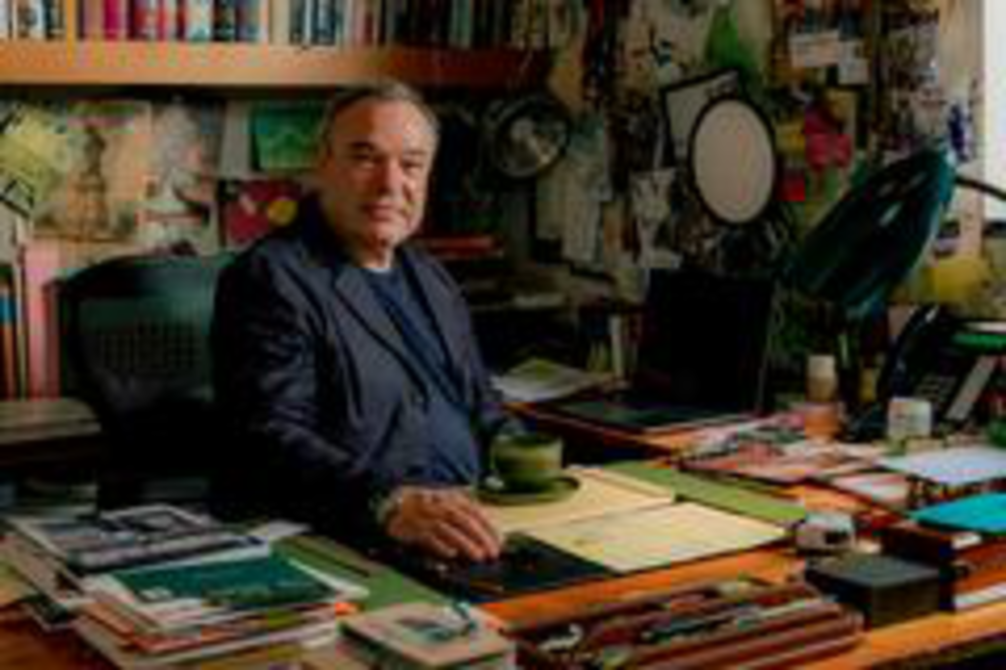 An older man sits at a cluttered wooden desk filled with papers and books, surrounded by bookshelves and leafy green plants in a cozy room.