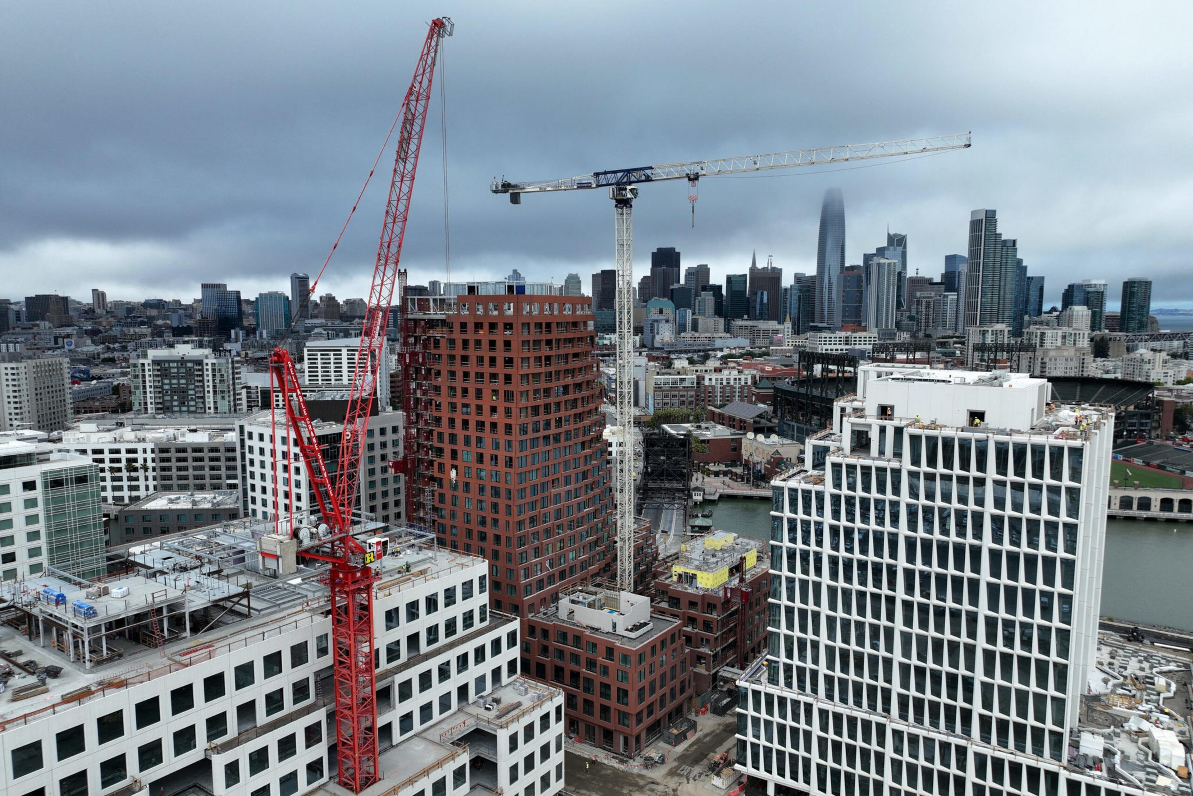 A city skyline with construction cranes, several mid-rise buildings in progress, and a river flowing alongside the urban area under a cloudy sky.