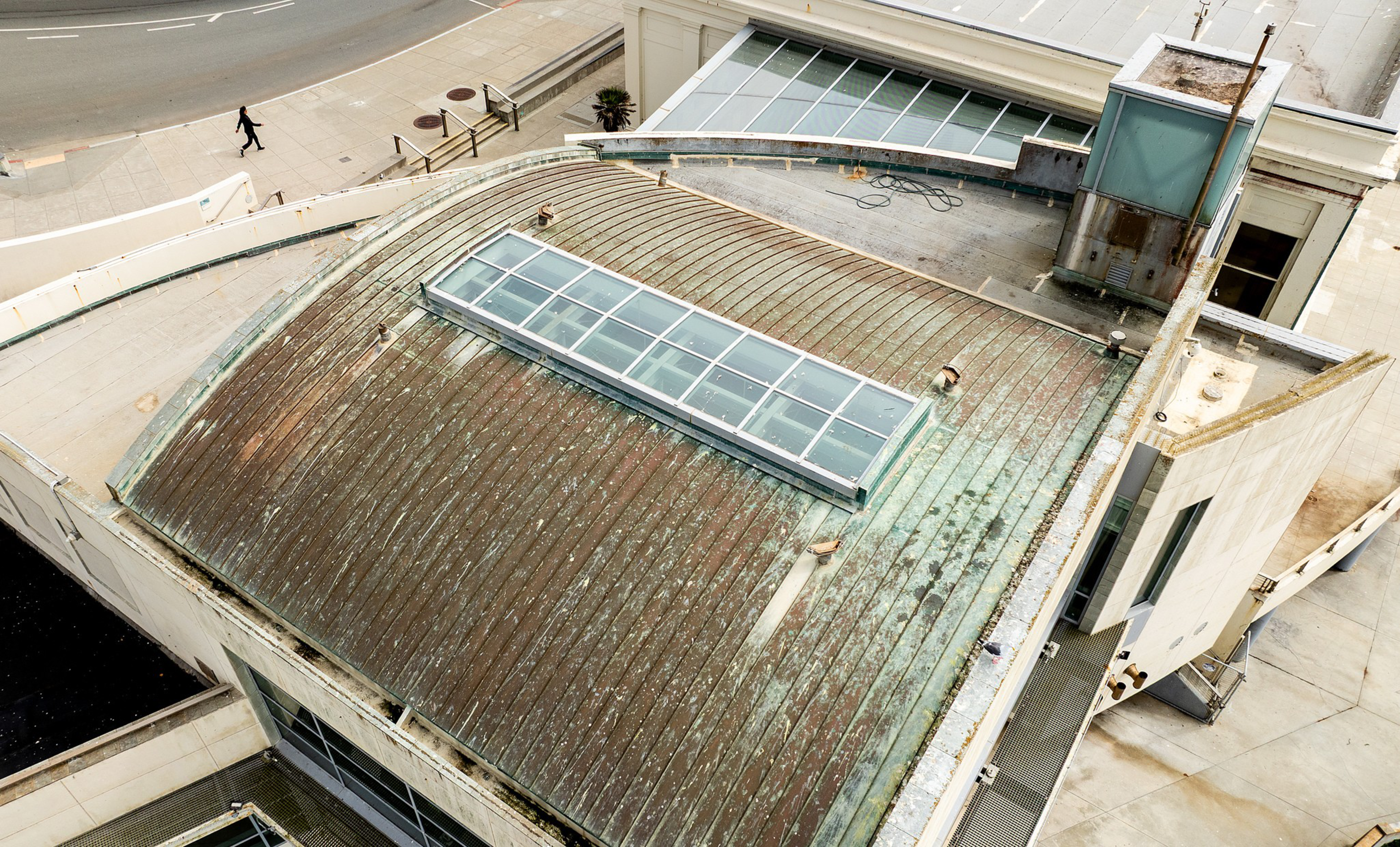 A weathered, curved metal rooftop with a long, narrow skylight in the center sits atop a white building near a street where a person walks.