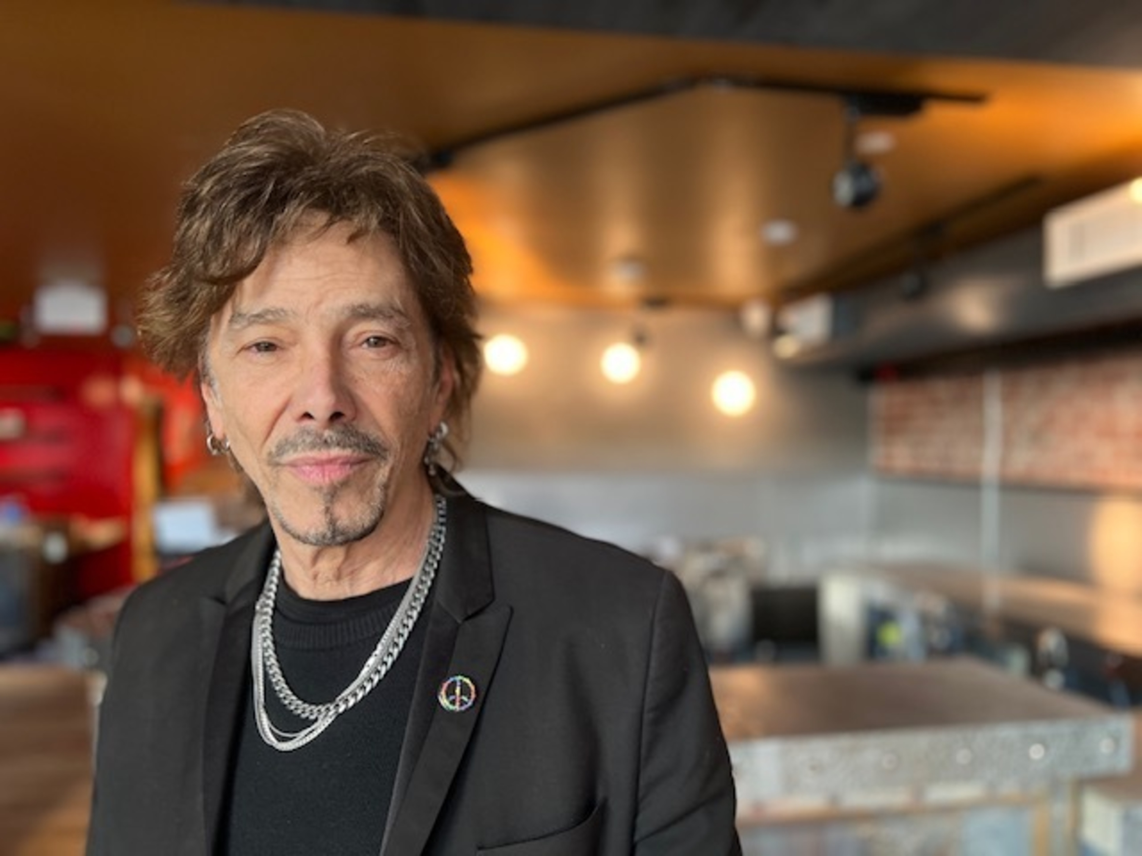 A man with silver chains and a black jacket stands in an industrial-looking space.
