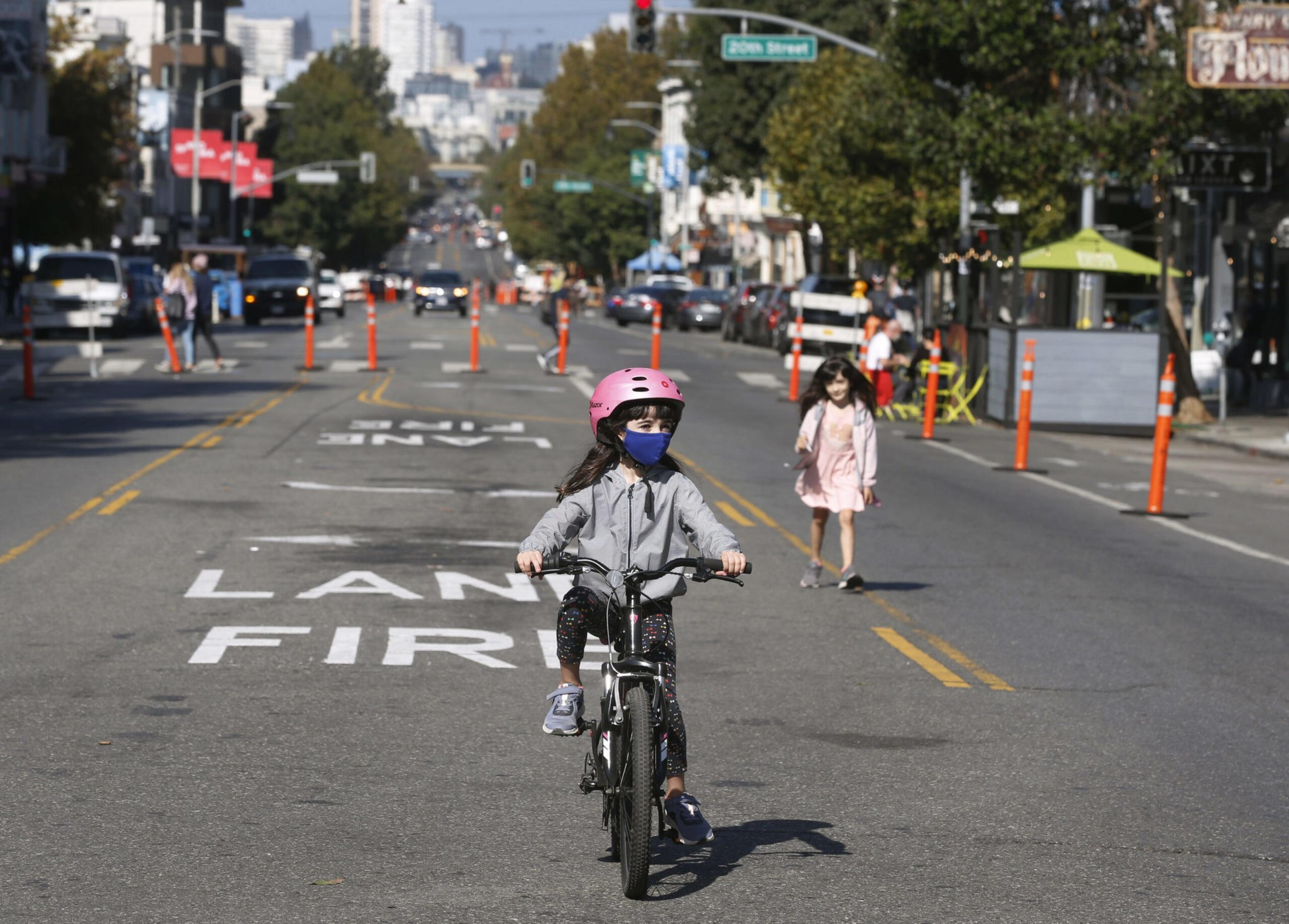 A child wearing a pink helmet and blue face mask rides a bike in the street, while another child in a pink dress walks behind, with cars and buildings in the background.