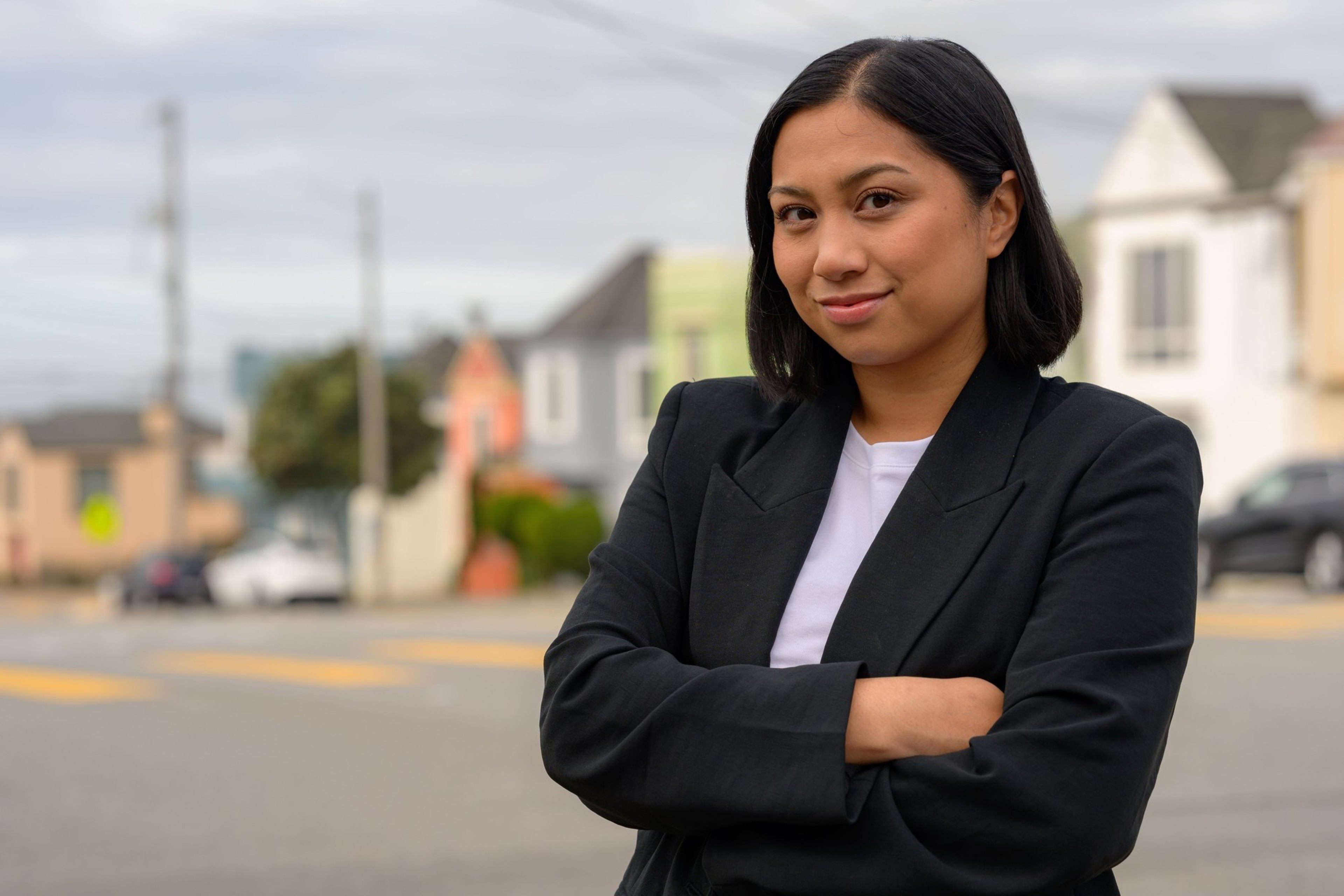 A young woman with shoulder-length black hair stands confidently with arms crossed, wearing a black blazer and white shirt, with houses and cars in the background.