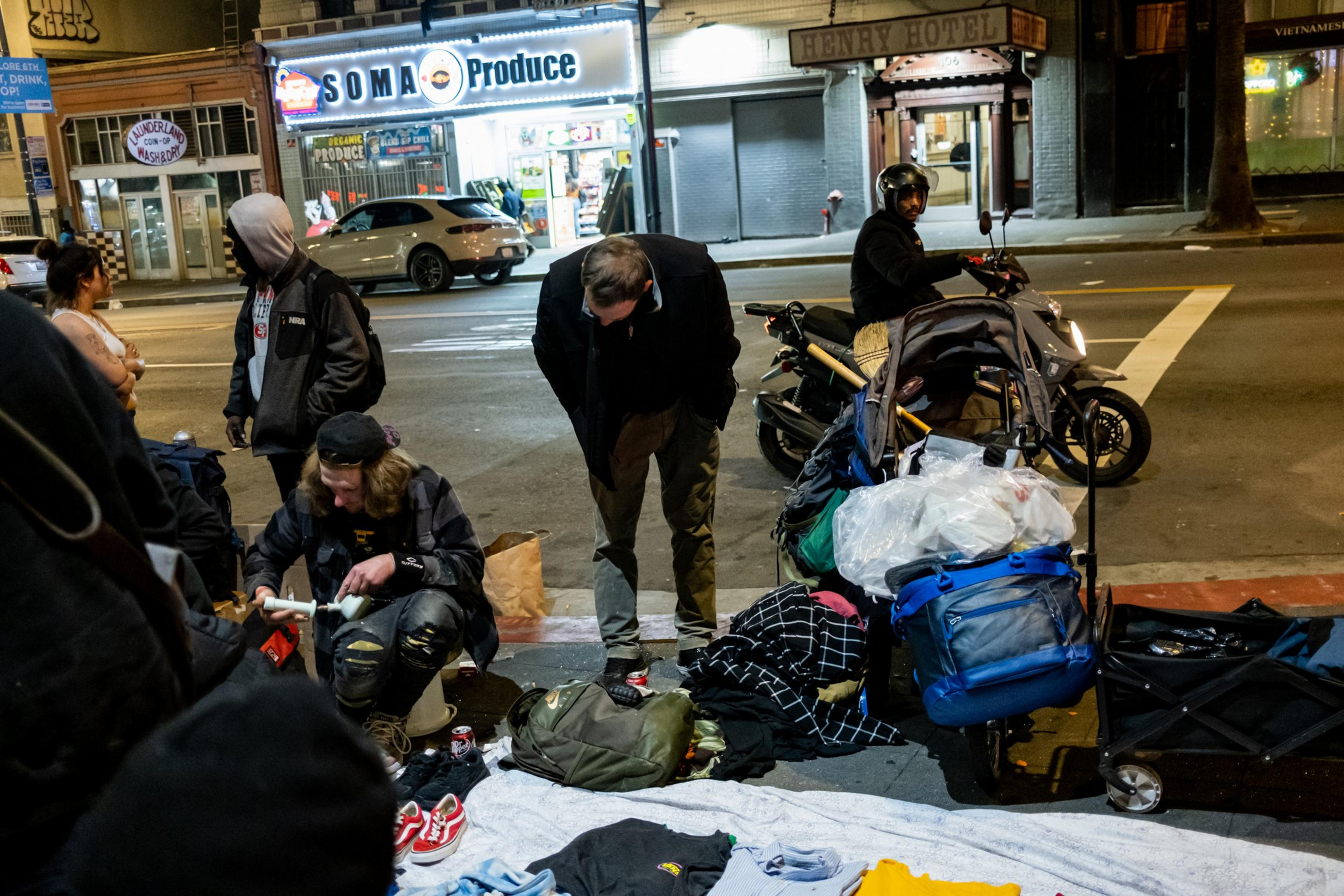 Several people stand and sit on a sidewalk near bags, clothing, and belongings at night, with a brightly lit store and a scooter on the street behind them.