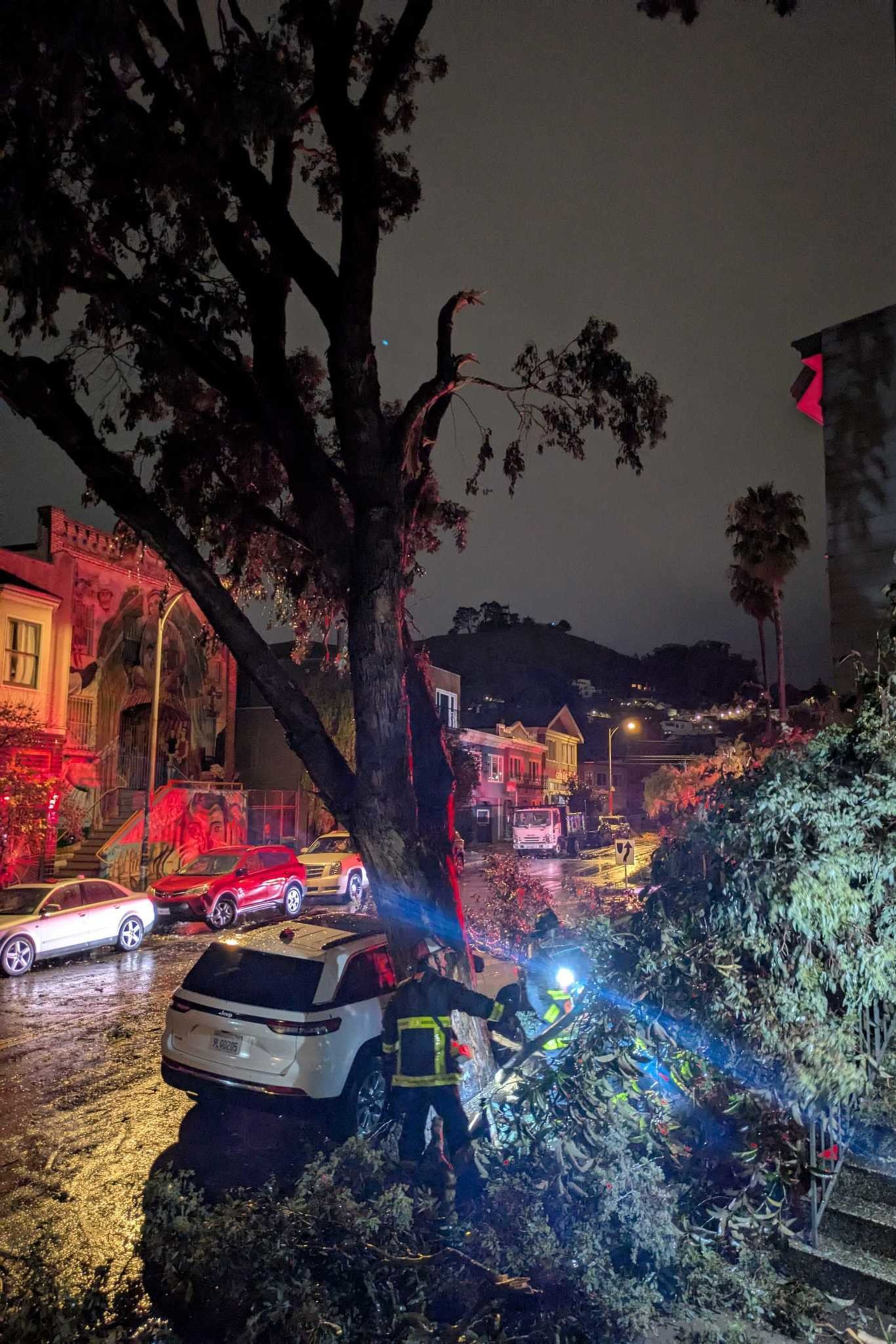 A large tree has fallen onto a white SUV on a dark, rainy street. Emergency workers in reflective gear are clearing the debris. Streetlights illuminate the scene.