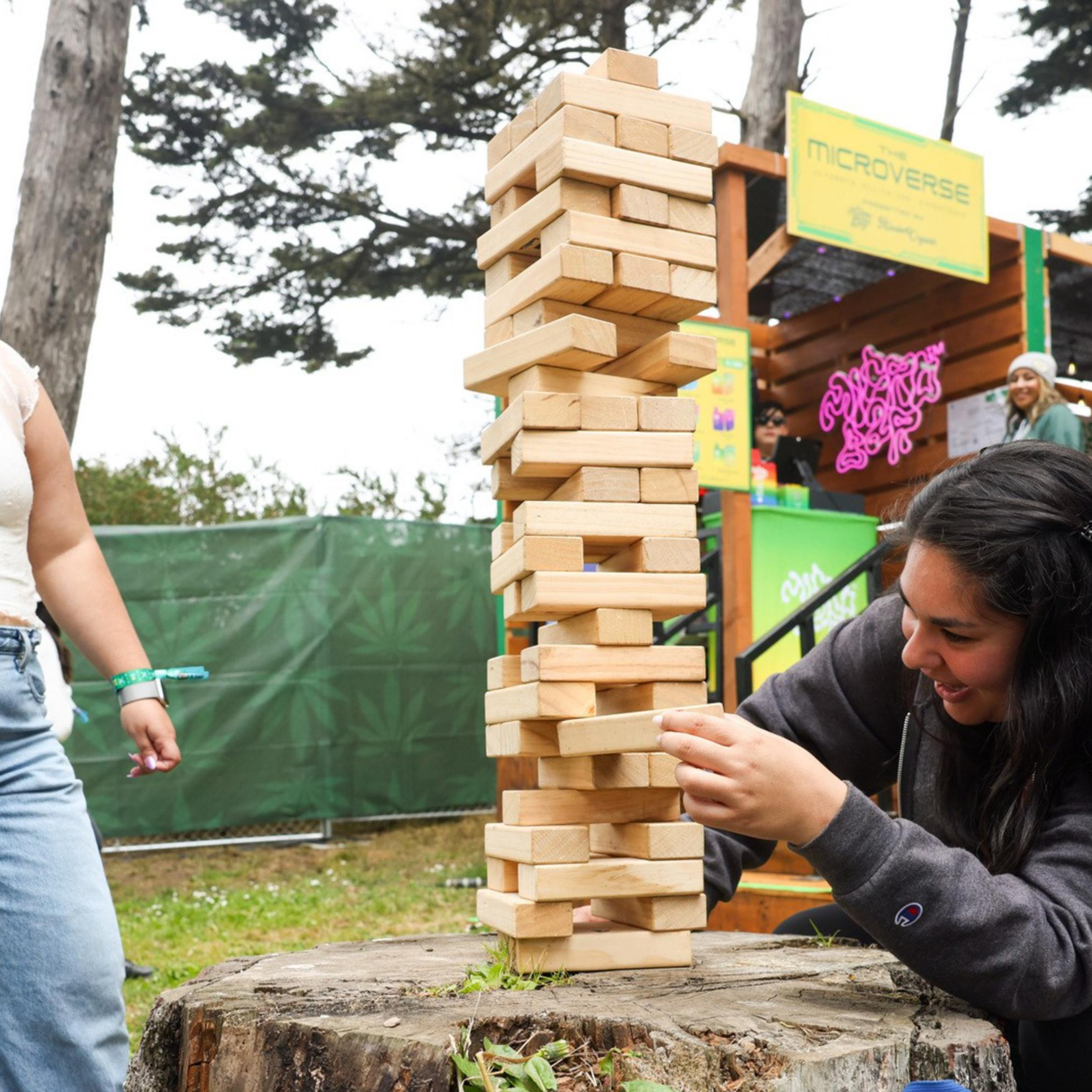 Two people enjoy a large Jenga game on a tree stump. One is pushing a block, while the other laughs. The scene is outdoors, near green fencing and booths.