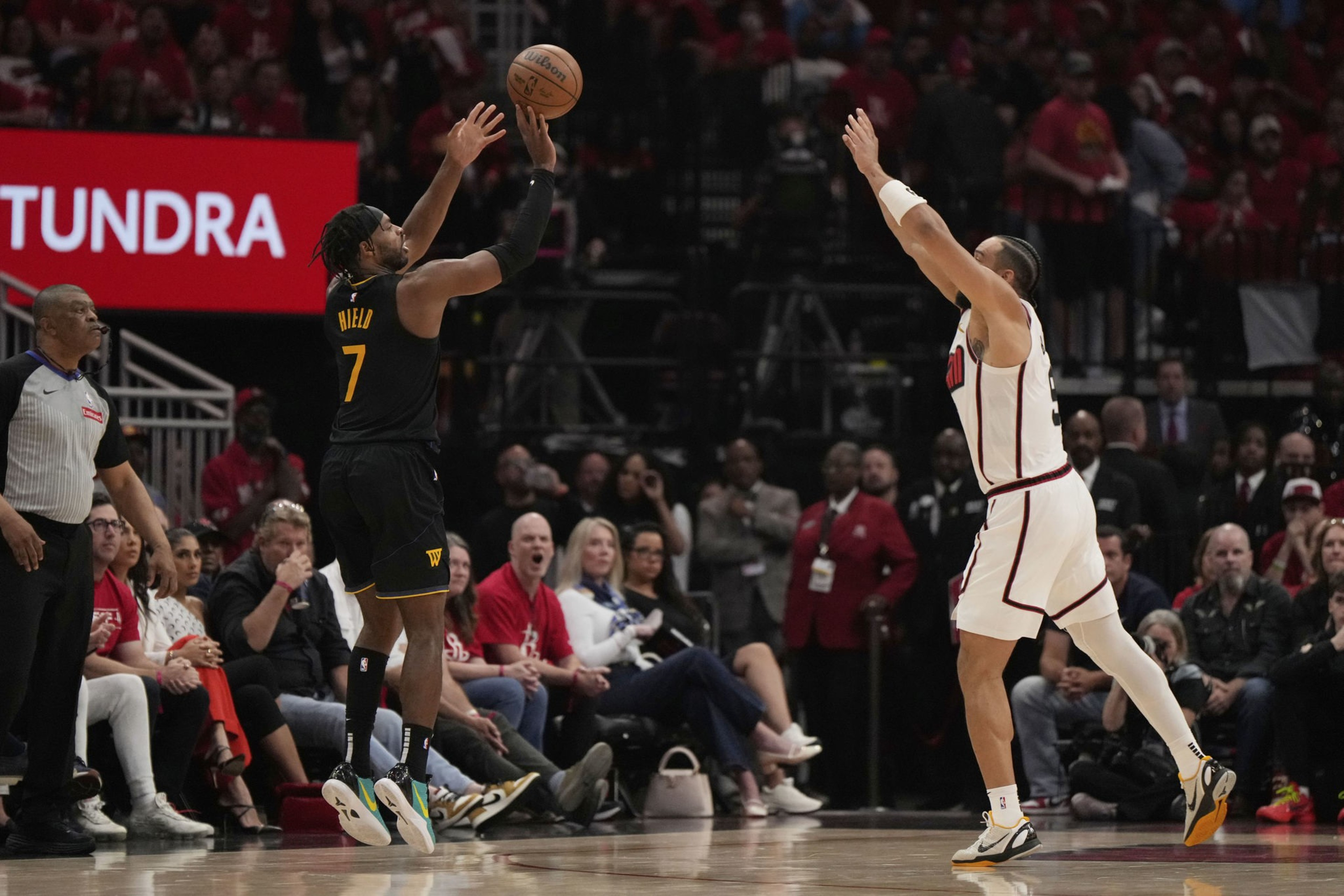 A basketball player in a black uniform jumps to shoot a ball, opposed by a player in a white uniform. A referee and spectators are visible in the background.