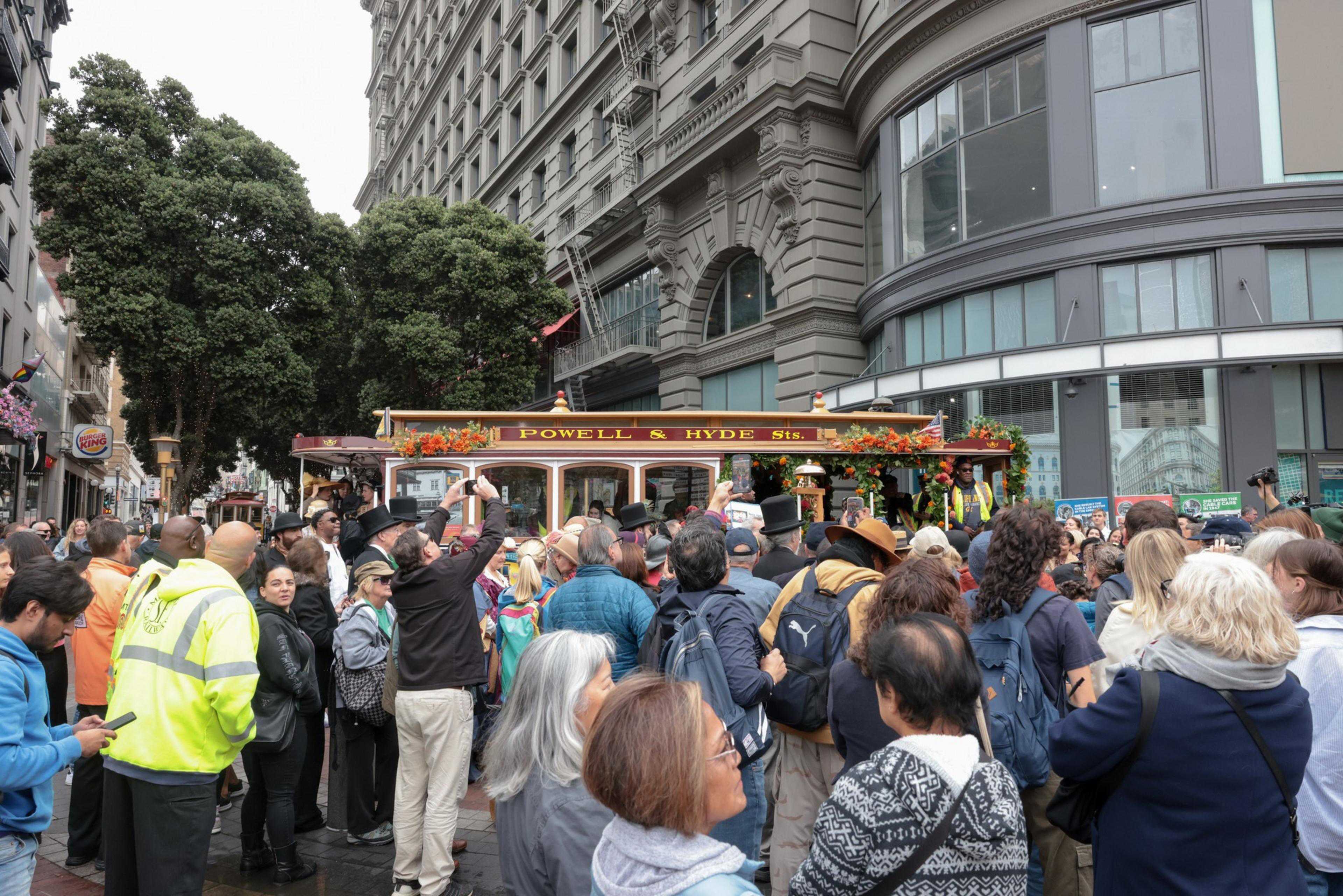 A crowded street scene shows people gathered around a cable car labeled "Powell & Hyde Sts." in an urban area with tall buildings and trees.