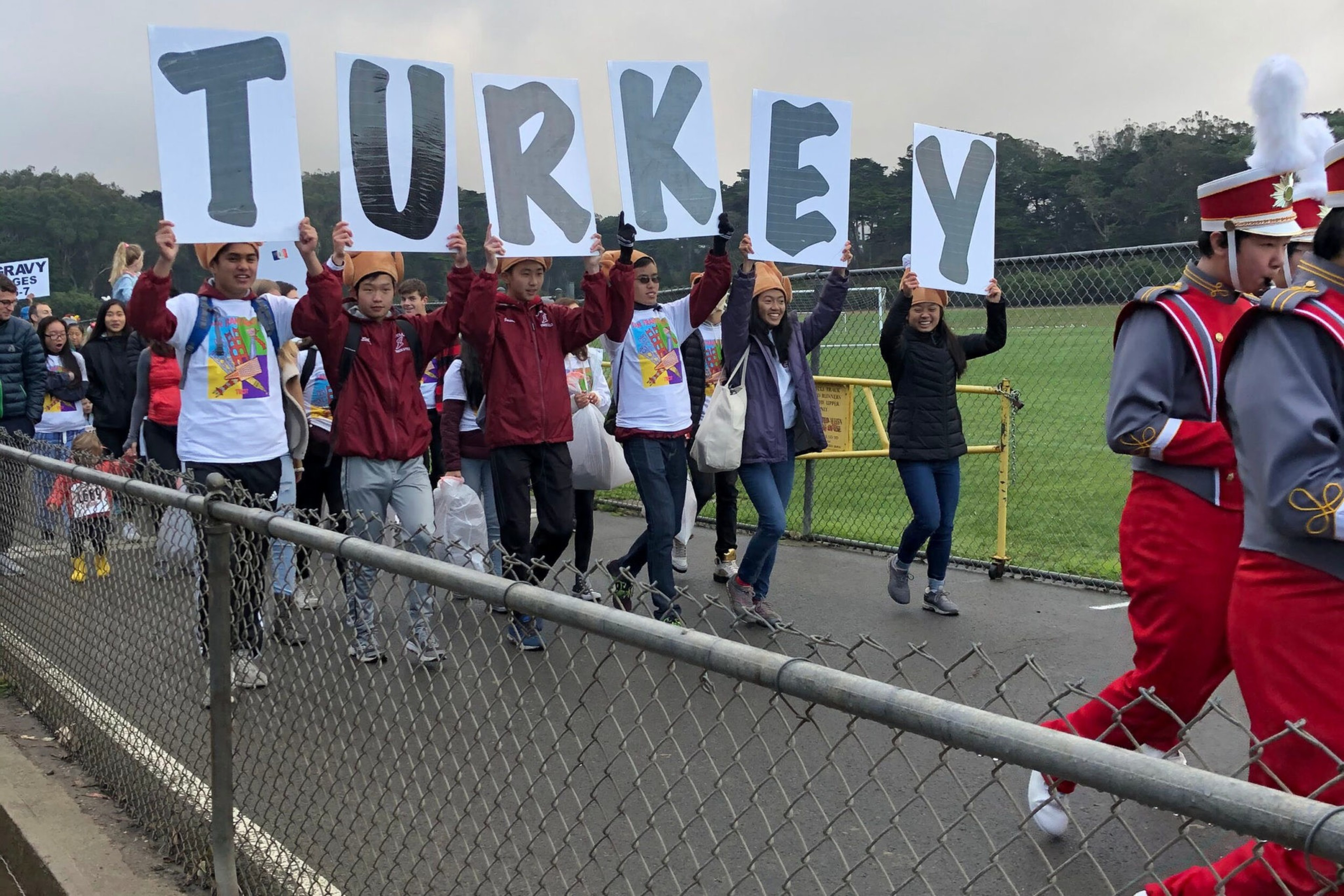 A group of people walk along a path holding signs that spell "TURKEY," while two marching band members in red and gray uniforms lead the way.