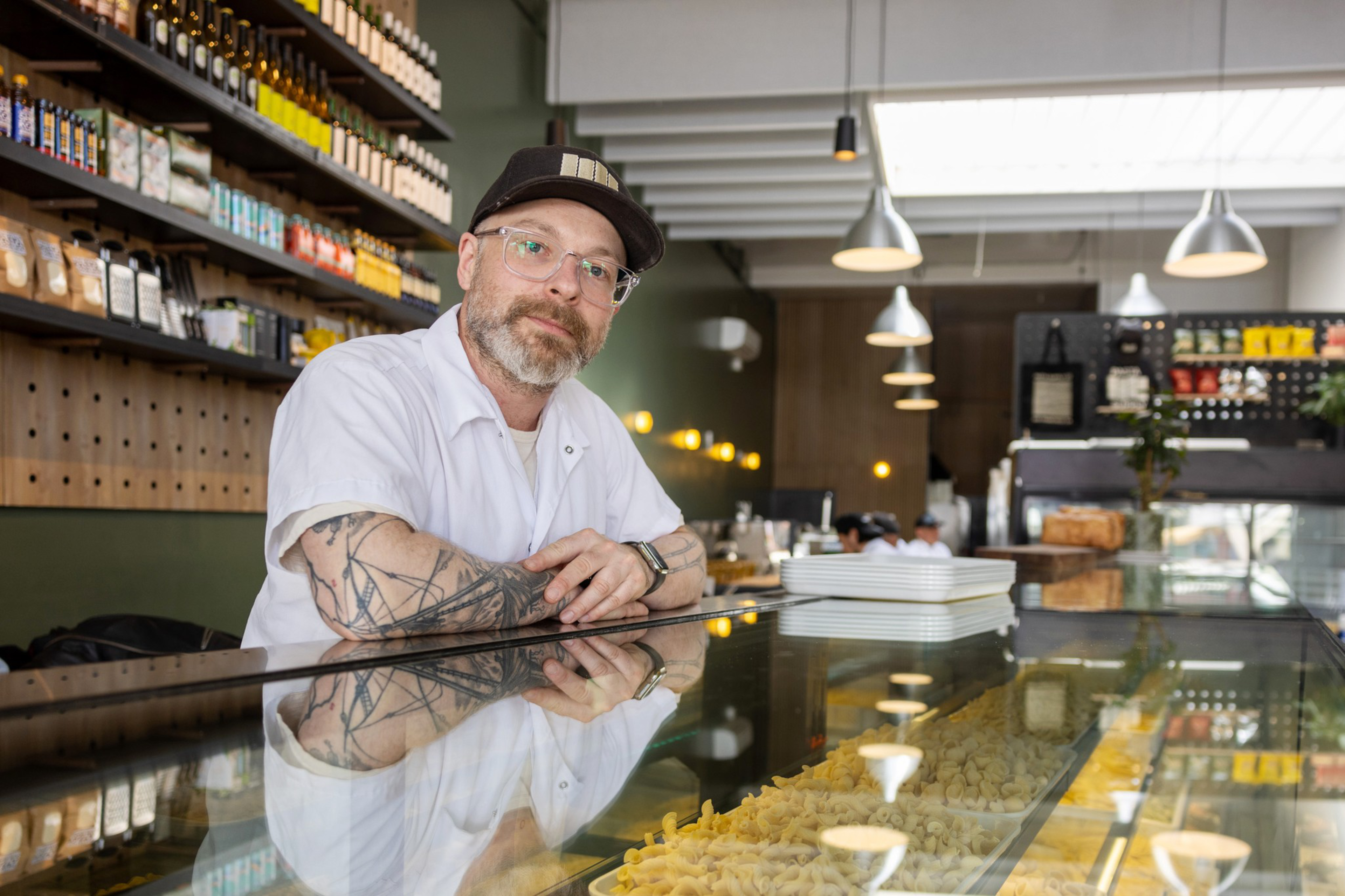 A man with tattoos and glasses, wearing a cap and a white uniform, leans on a glass counter filled with pasta in a well-lit store with shelves of products behind him.