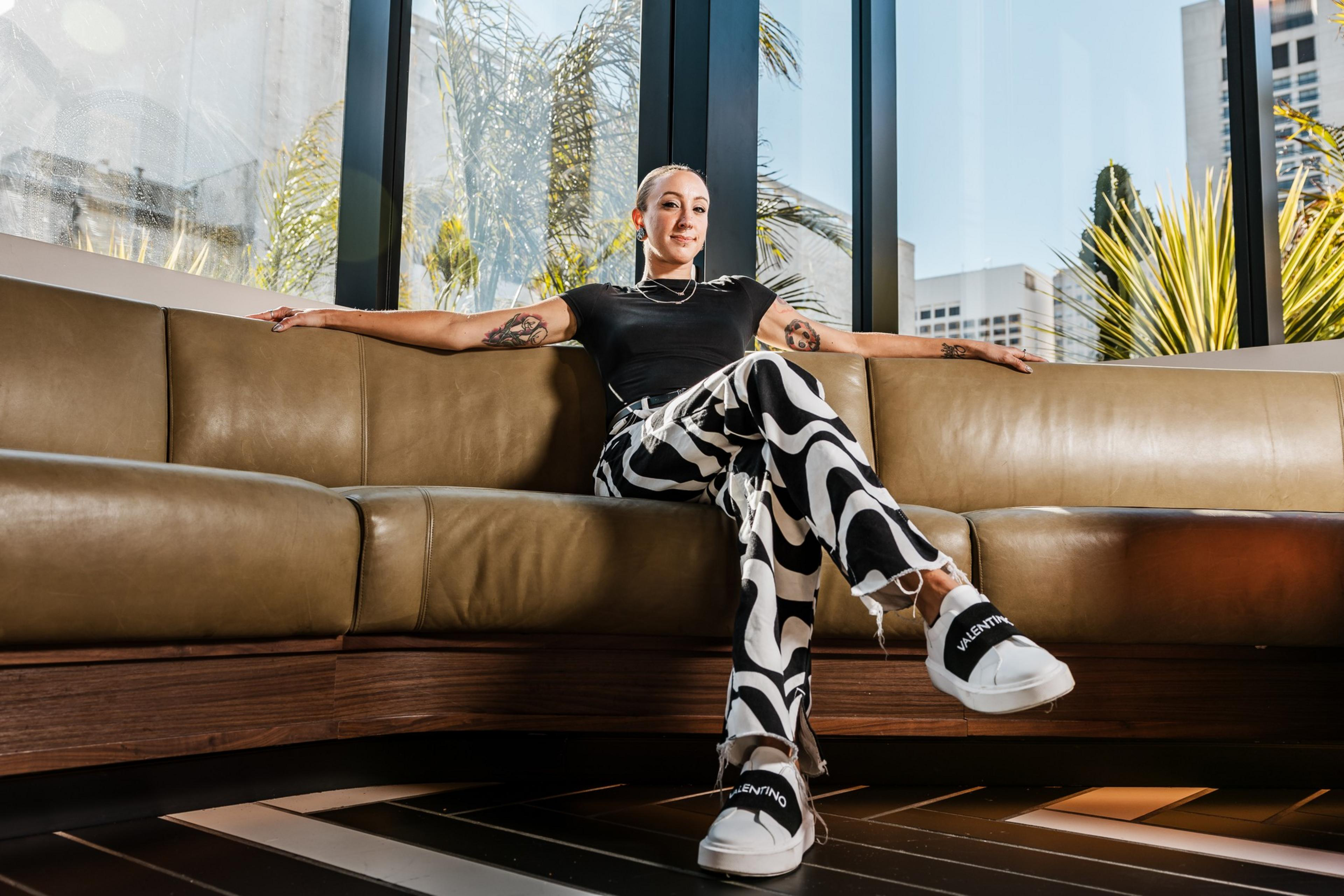 A person with a shaved head wearing a black shirt and patterned pants sits relaxed on a leather couch in a bright, modern room with large windows and plants outside.