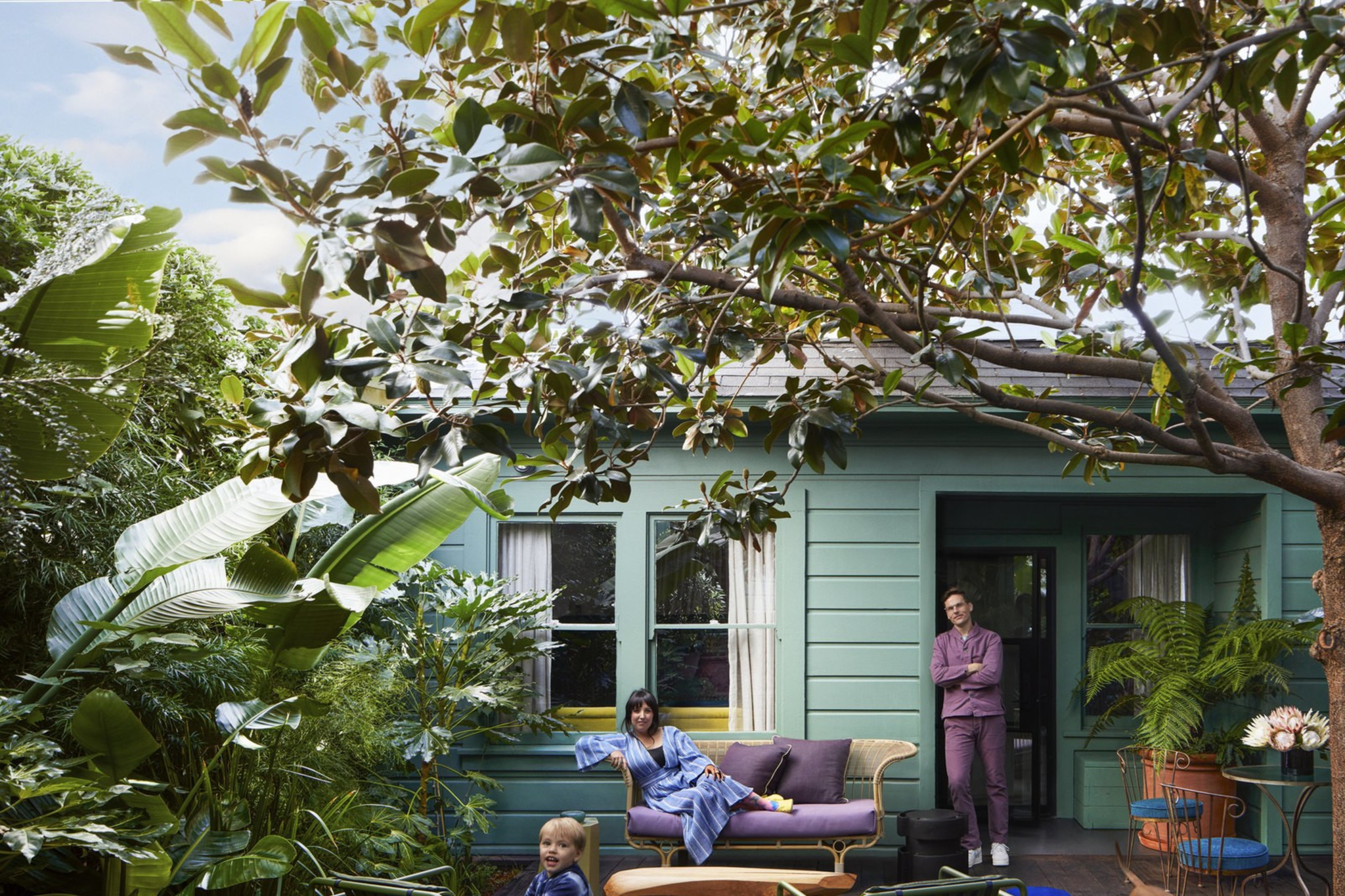 A family relaxes on a green-painted porch surrounded by lush plants, with a child playing, a woman seated, a man leaning by the door, and a dog walking nearby.