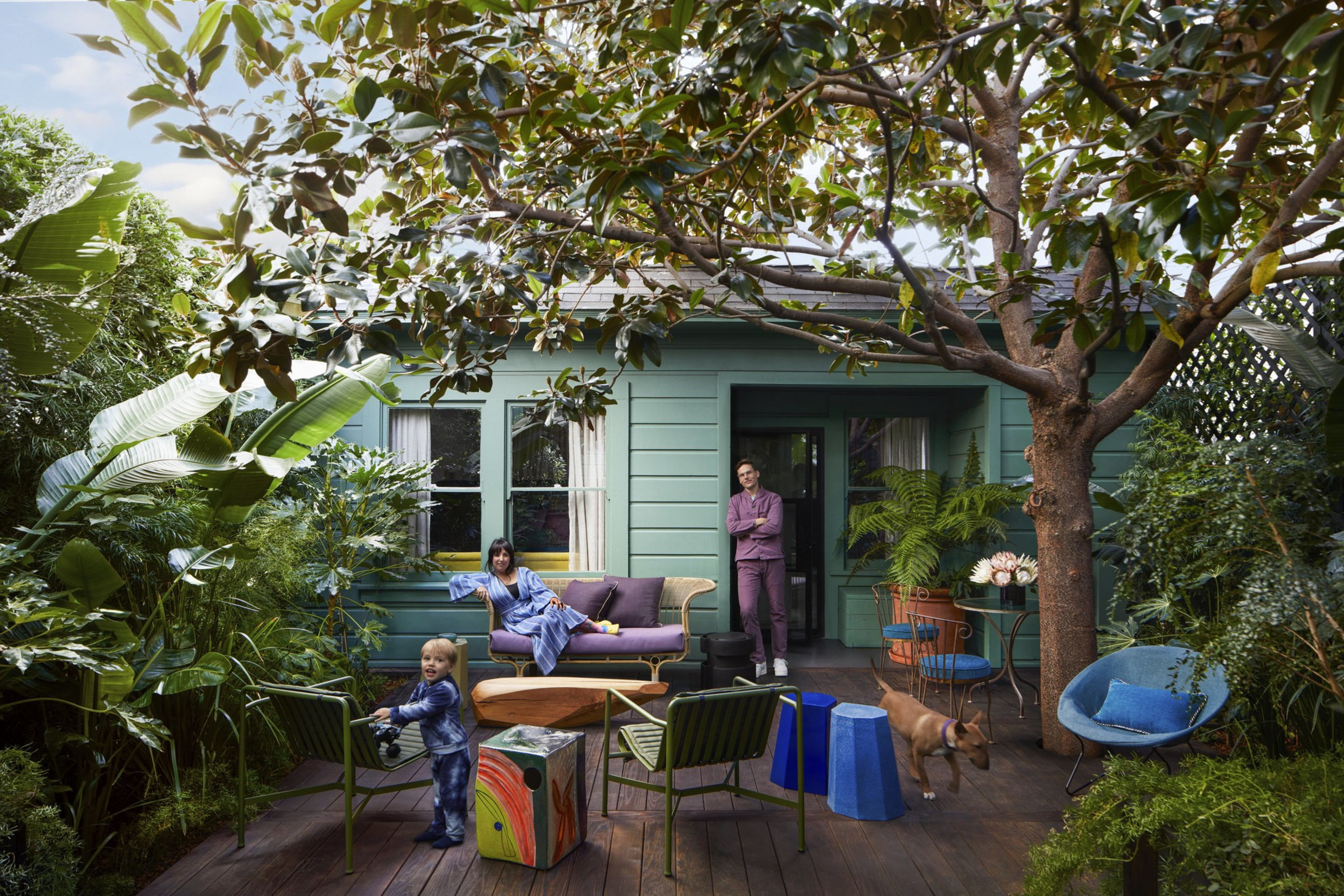 A family relaxes on a green-painted porch surrounded by lush plants, with a child playing, a woman seated, a man leaning by the door, and a dog walking nearby.