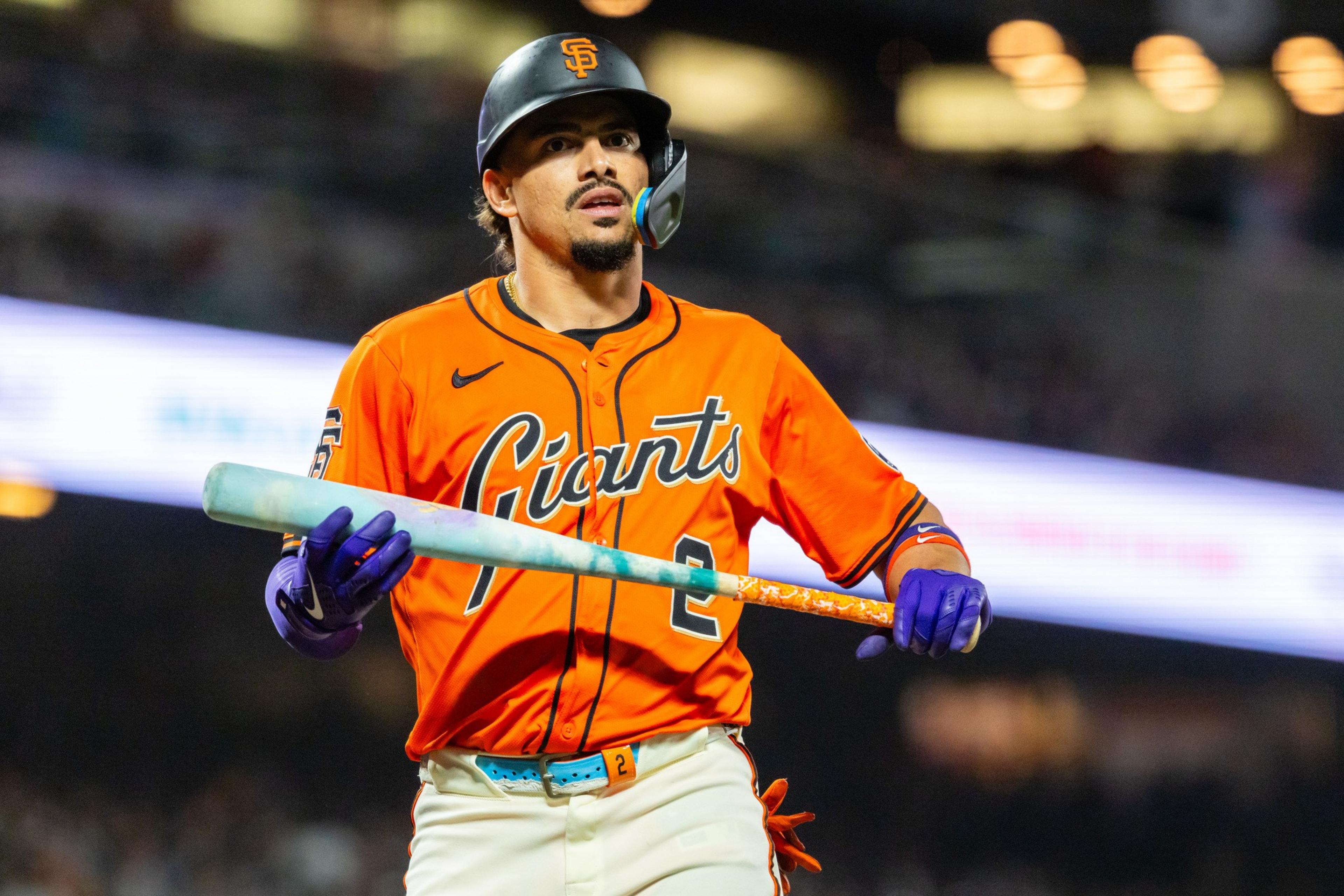 A baseball player in an orange Giants jersey and black helmet holds a bat with purple gloves, looking focused during a night game.