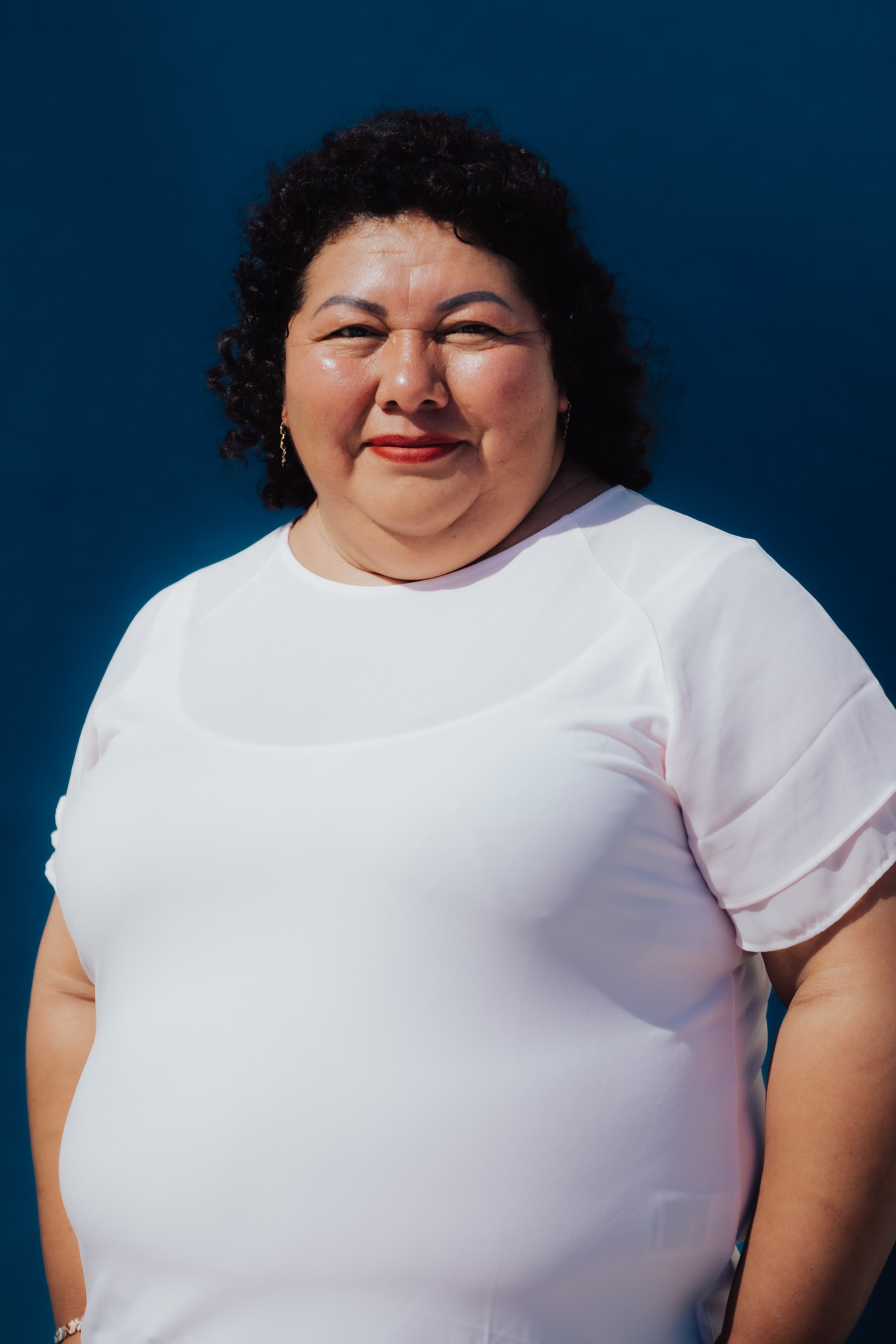 A smiling woman with curly dark hair wears a white shirt and gold earrings, standing against a solid dark blue background.