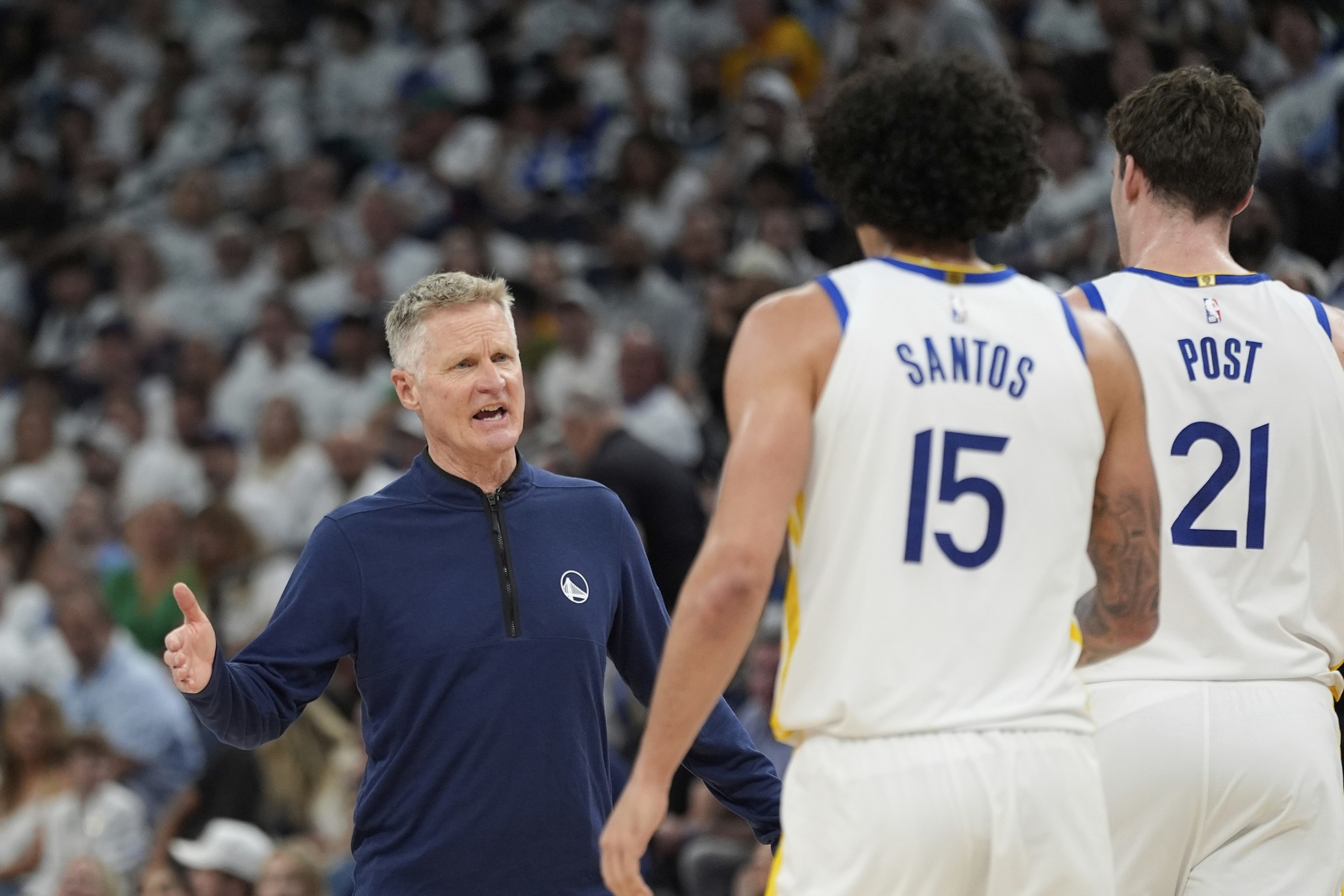 A coach gestures while talking to two basketball players wearing white jerseys numbered 15 and 21. The background shows a crowd in a sports arena.