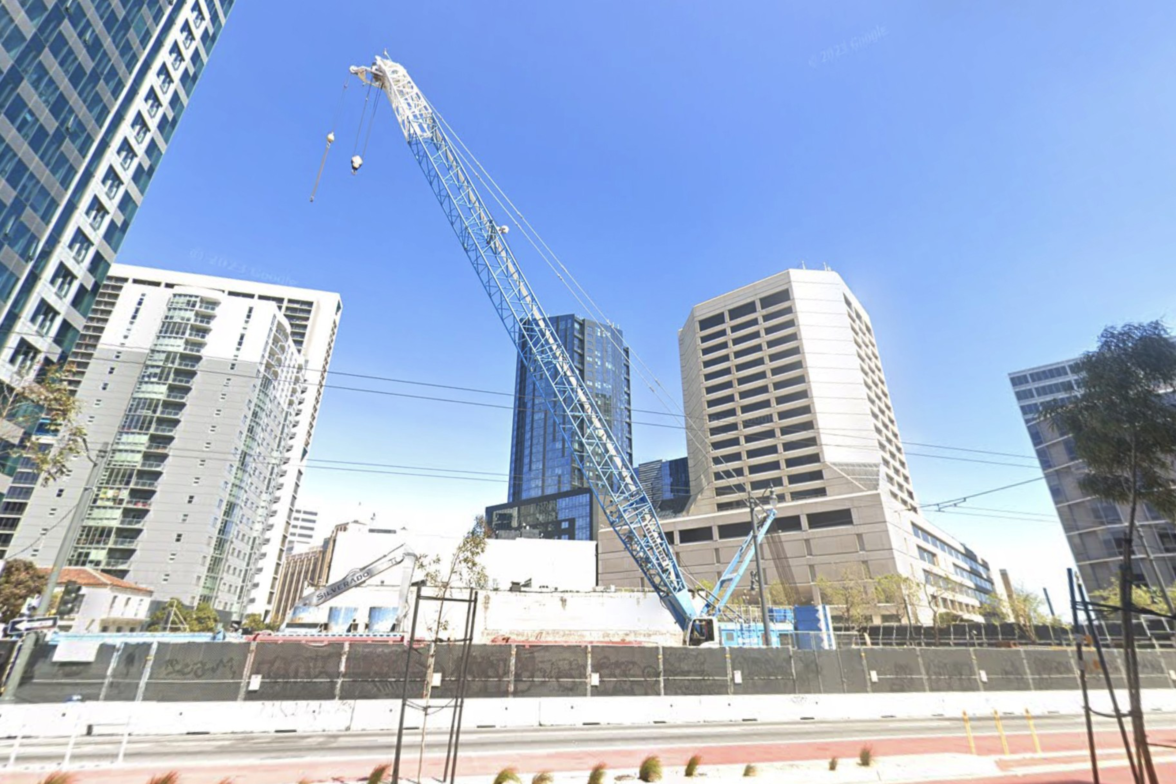 A large construction crane towers over a fenced construction site surrounded by tall office buildings under a clear blue sky.