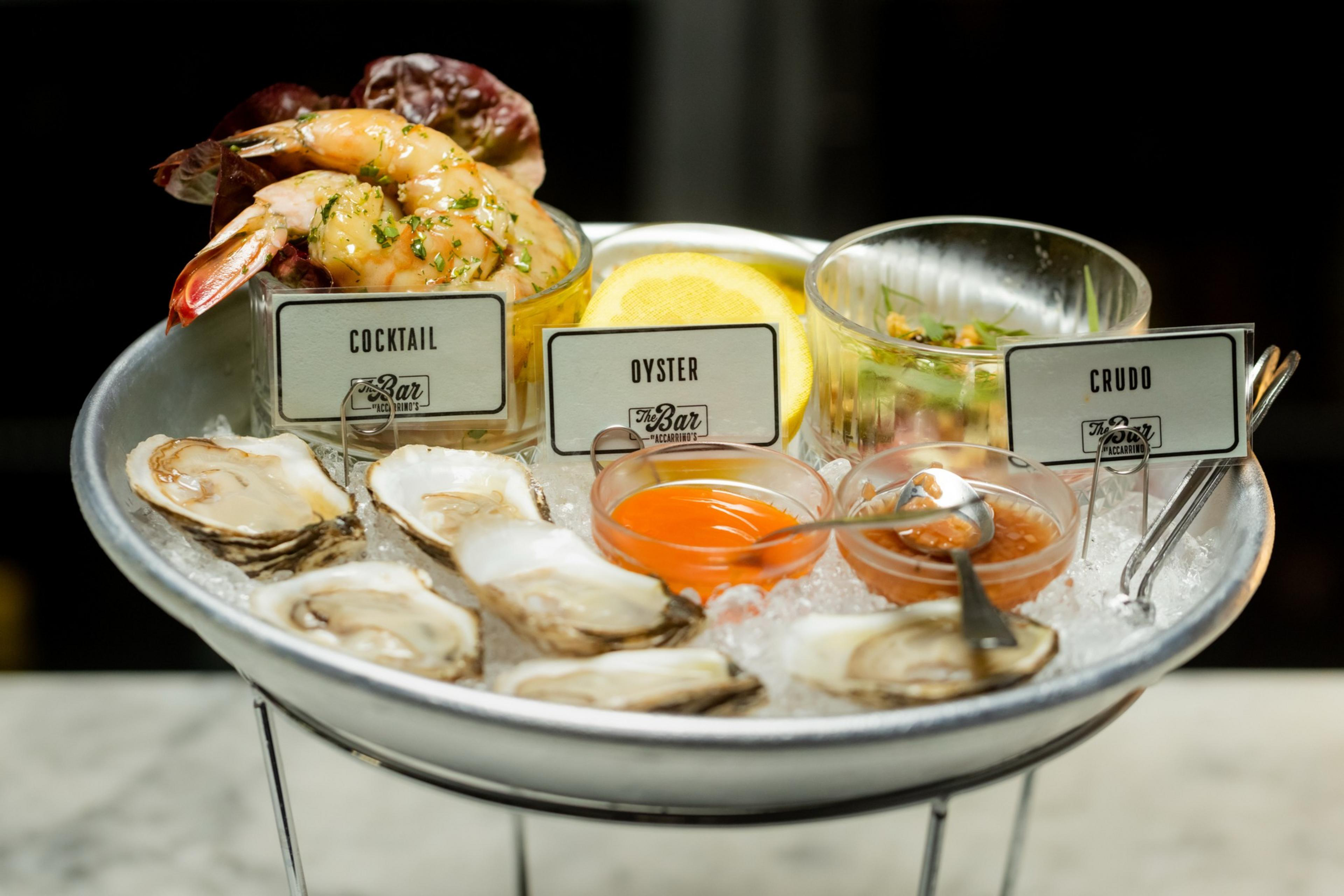 A seafood platter with shrimp cocktail, oysters, and a crudo dish, served on ice alongside lemon slices and two small sauces in bowls.