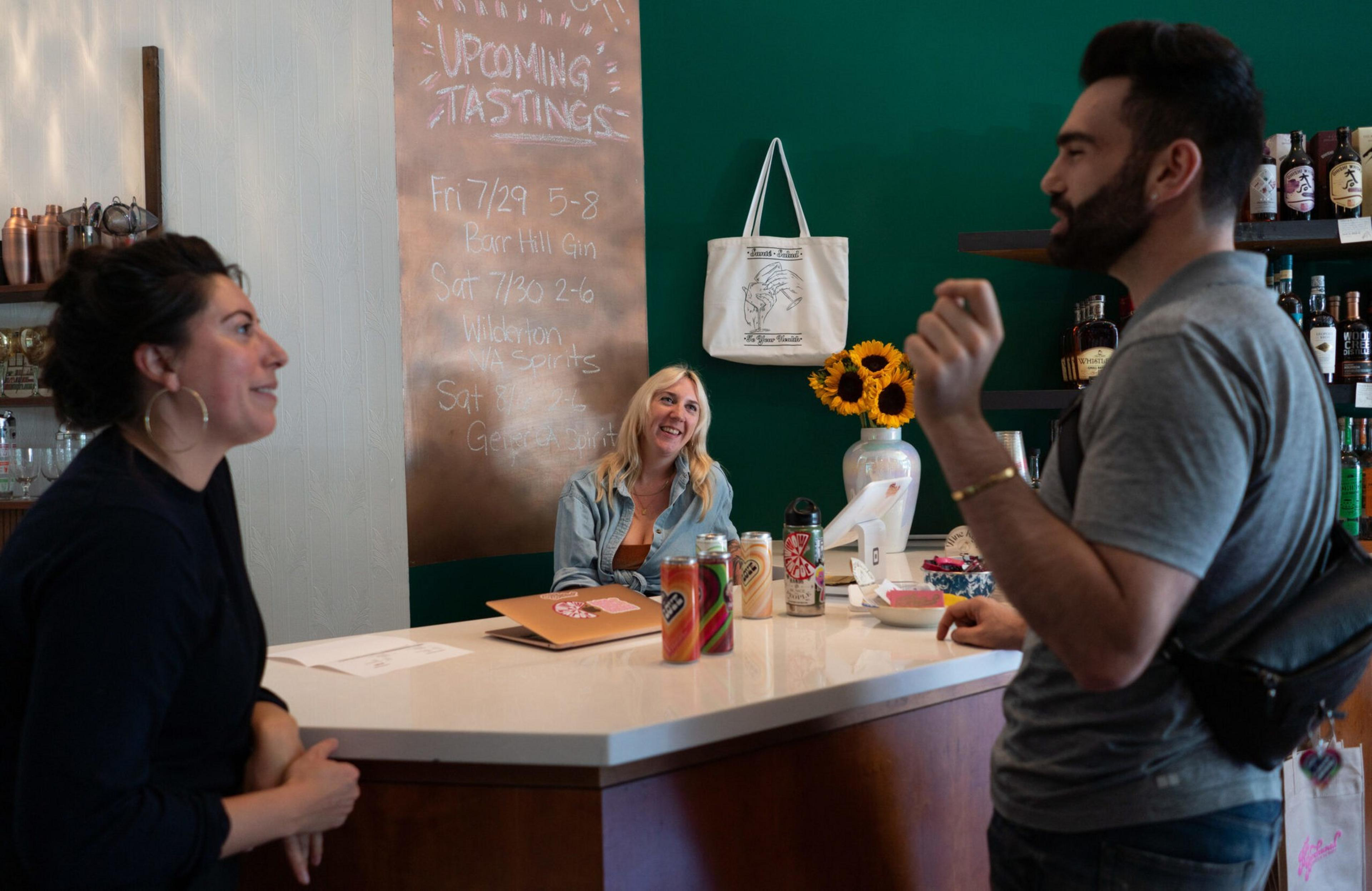 Three people are conversing at a bar counter with cans and bottles on it, a chalkboard listing upcoming tastings on the wall behind them.