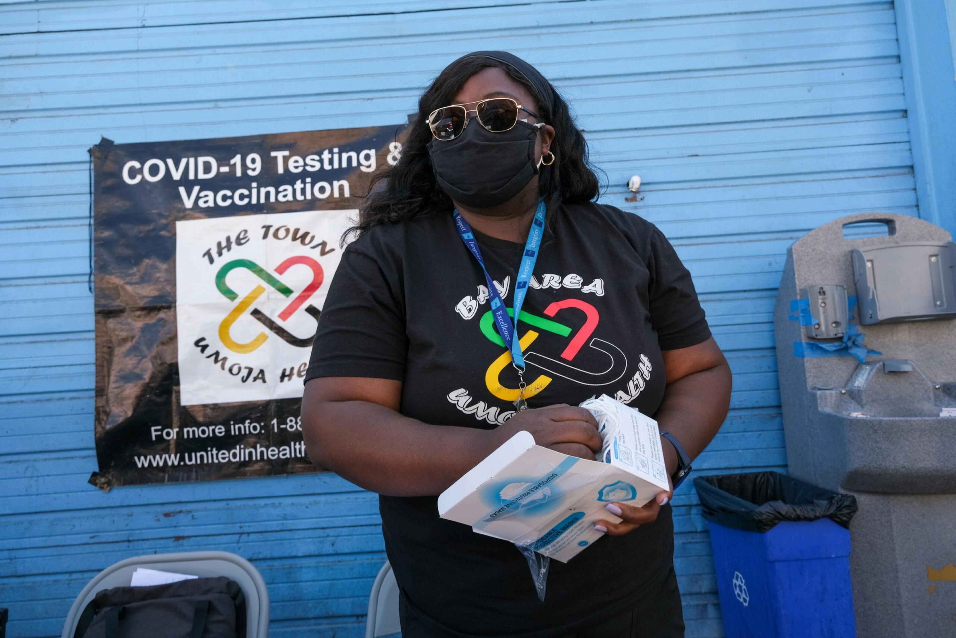 A person wearing sunglasses and a black mask holds a box of disposable gloves in front of a blue wall with a COVID-19 testing and vaccination sign.