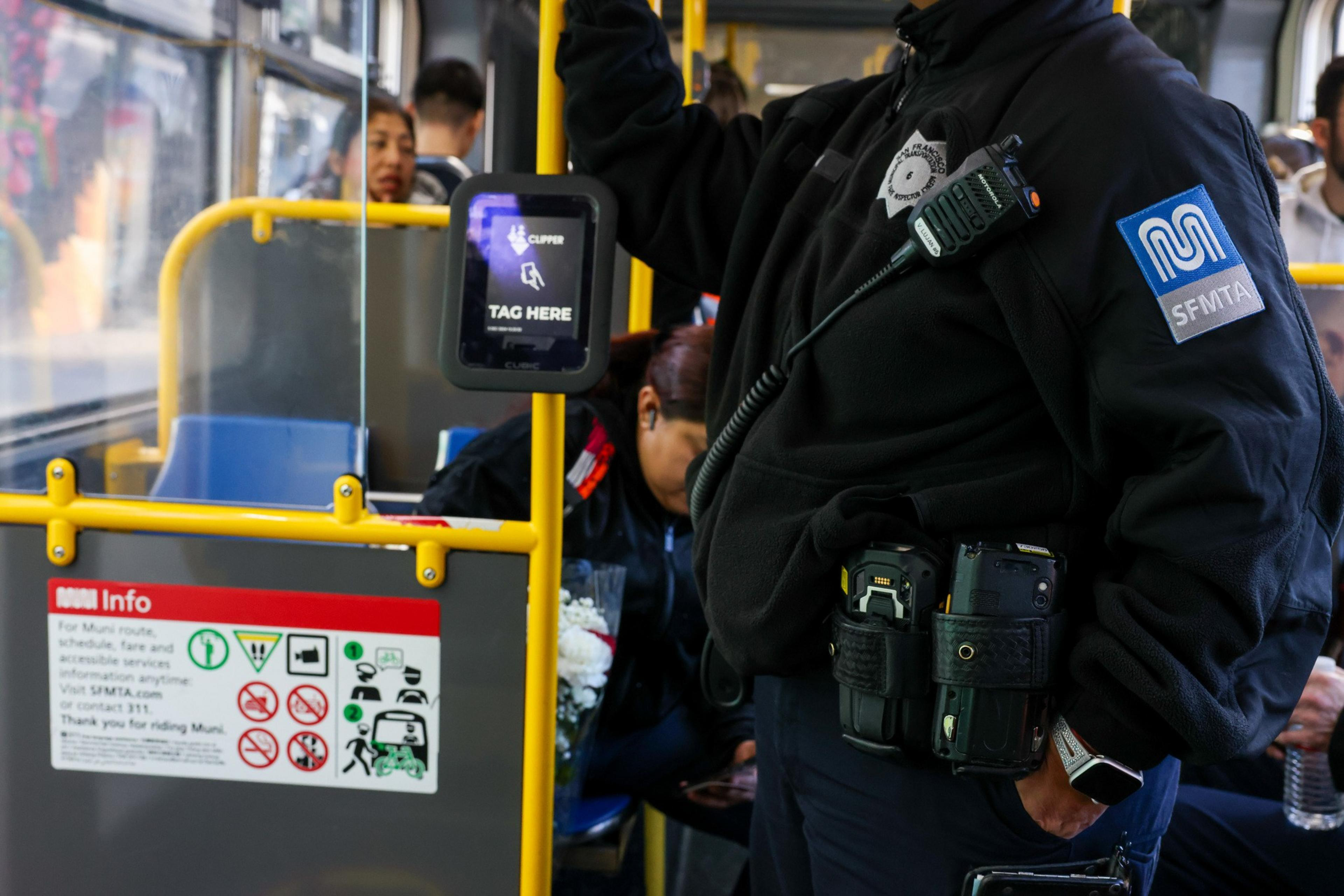 A transportation officer with SFMTA gear stands on a bus. Nearby are passengers seated, a Clipper card reader, and a sign with transit info and rules.