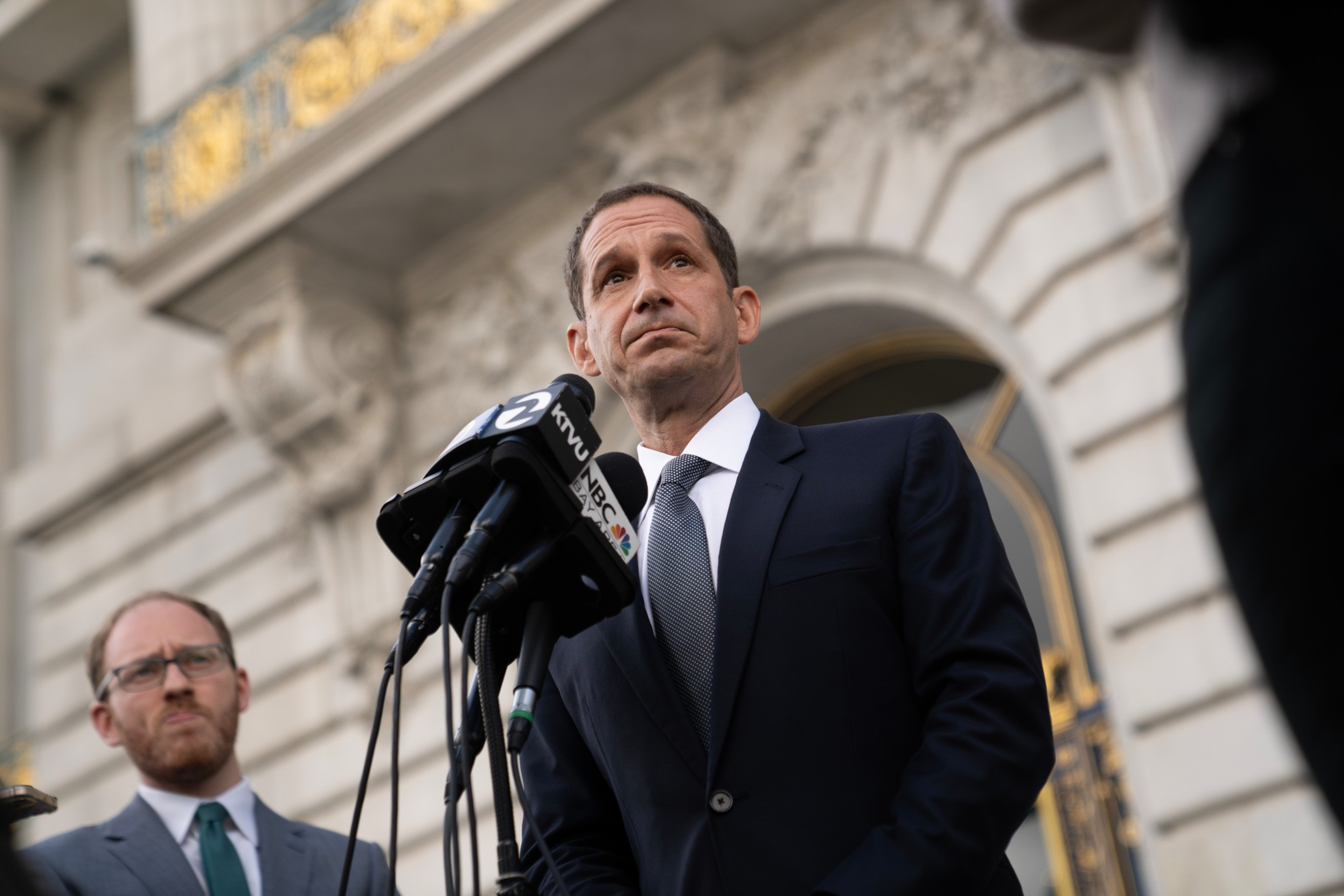 A man in a suit speaks seriously at microphones labeled KTVU and ABC, with another man in a suit standing behind him outside a stone building.