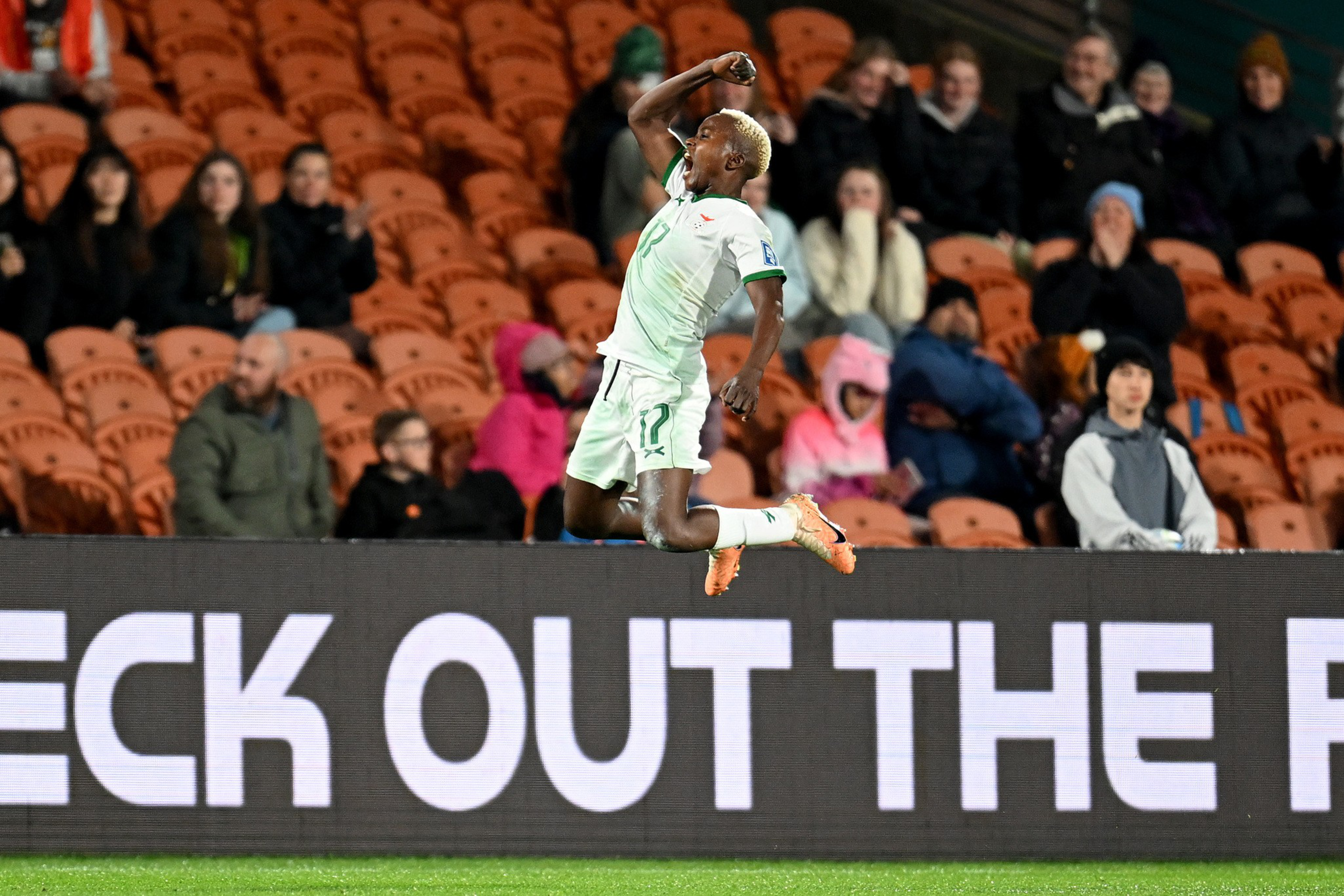 A soccer player in a white and green uniform jumps in celebration with a clenched fist near a digital advertising board, with seated spectators behind.