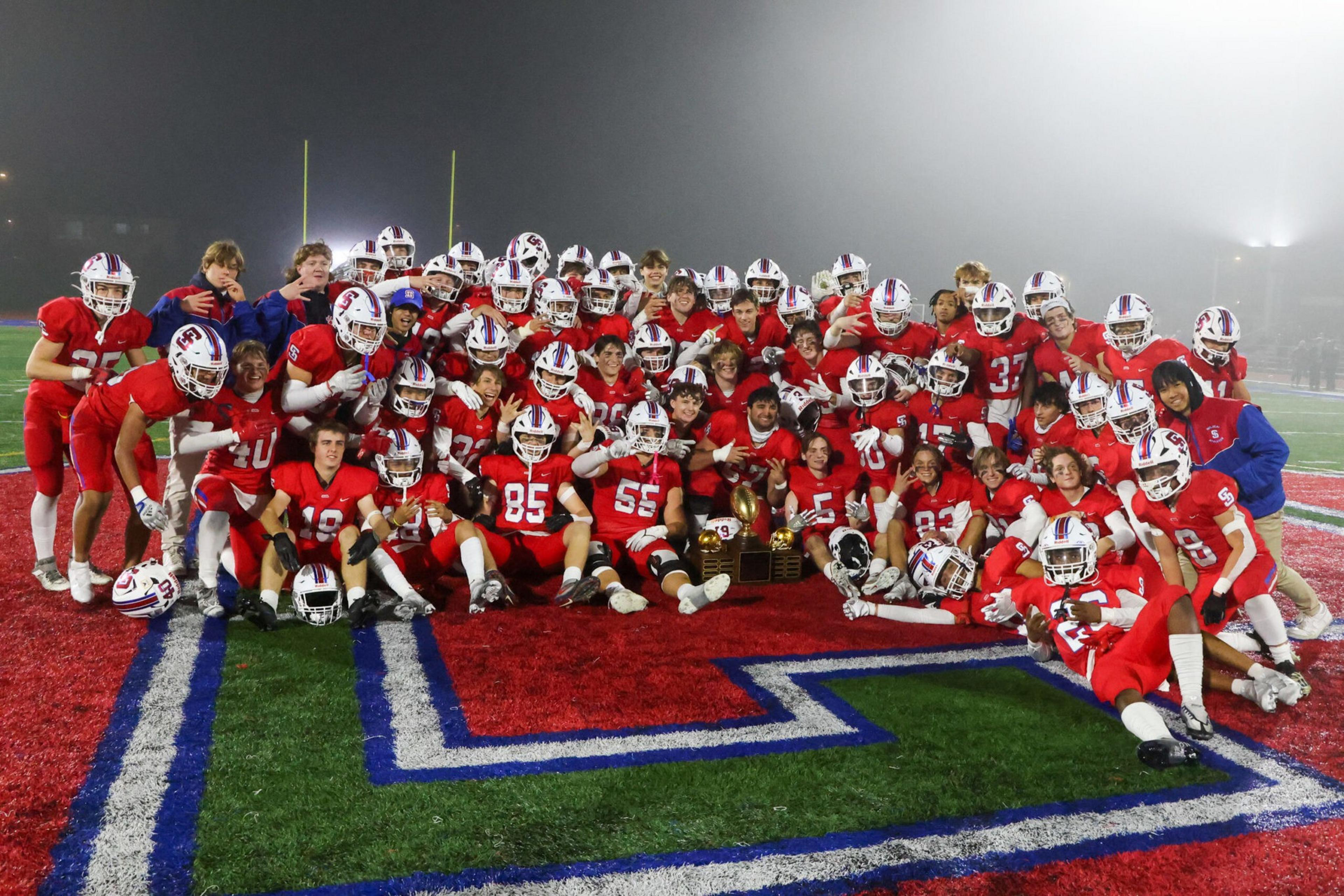 A football team in red uniforms poses on a field's large red and blue logo, holding a trophy and celebrating together.