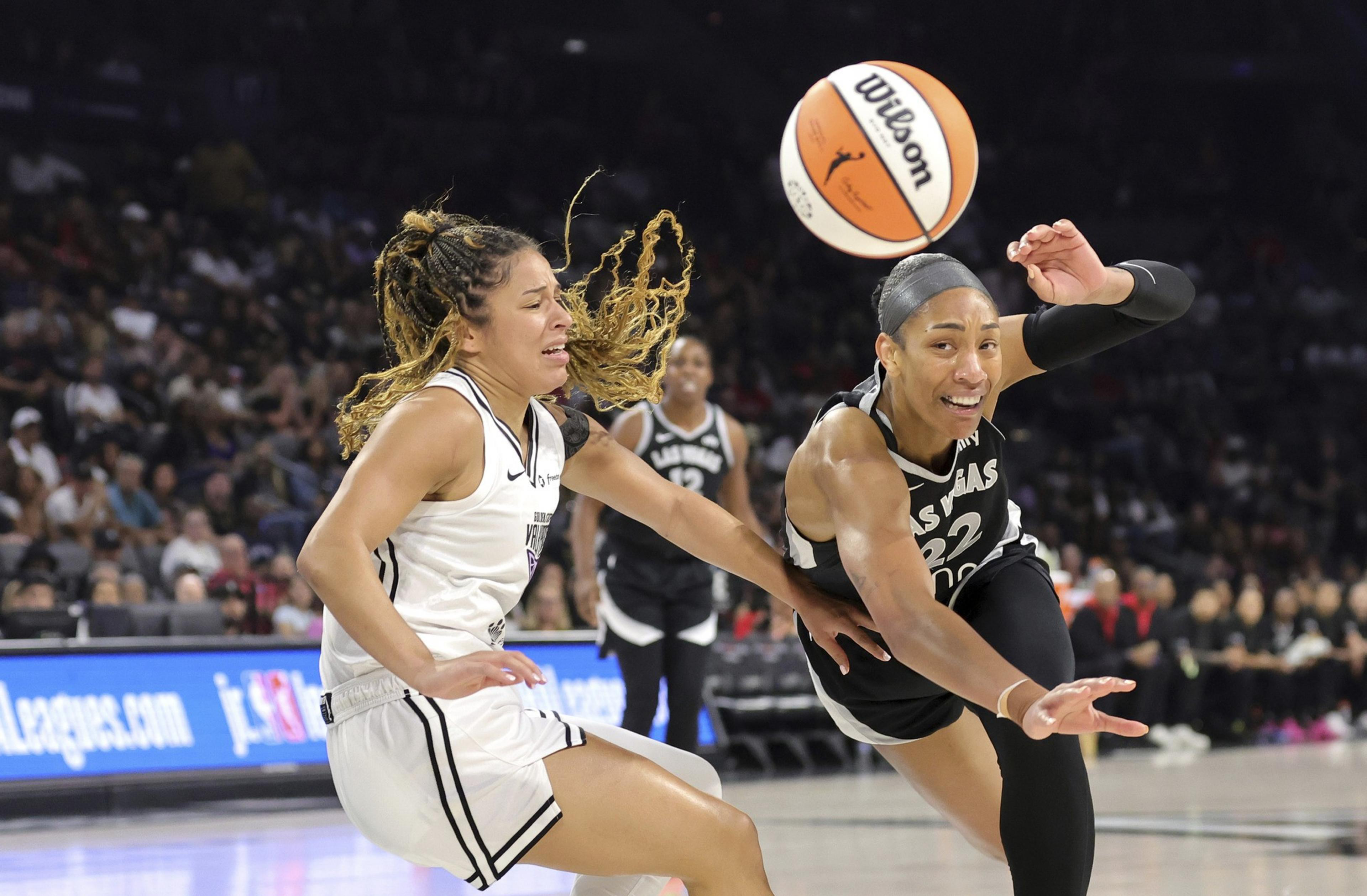 Two female basketball players in intense action, one reaching for the ball mid-air while the other grimaces, with a crowd watching in the background.