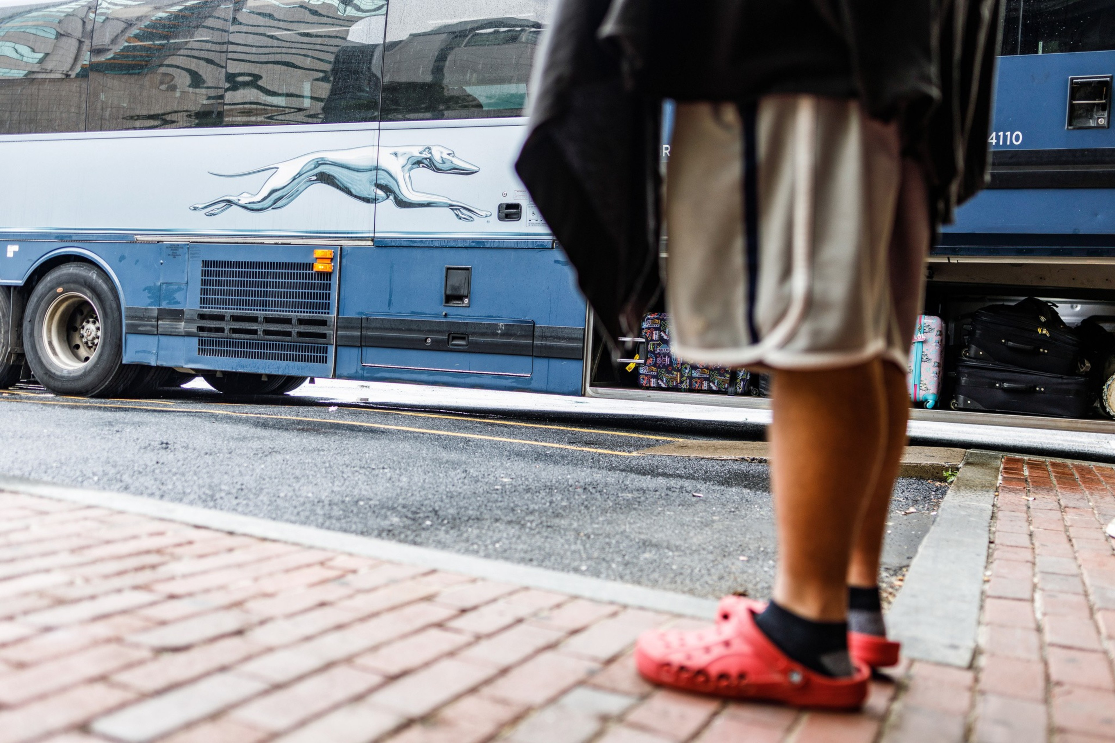 A person in red shoes stands by a blue bus with a greyhound logo; luggage is visible underneath.