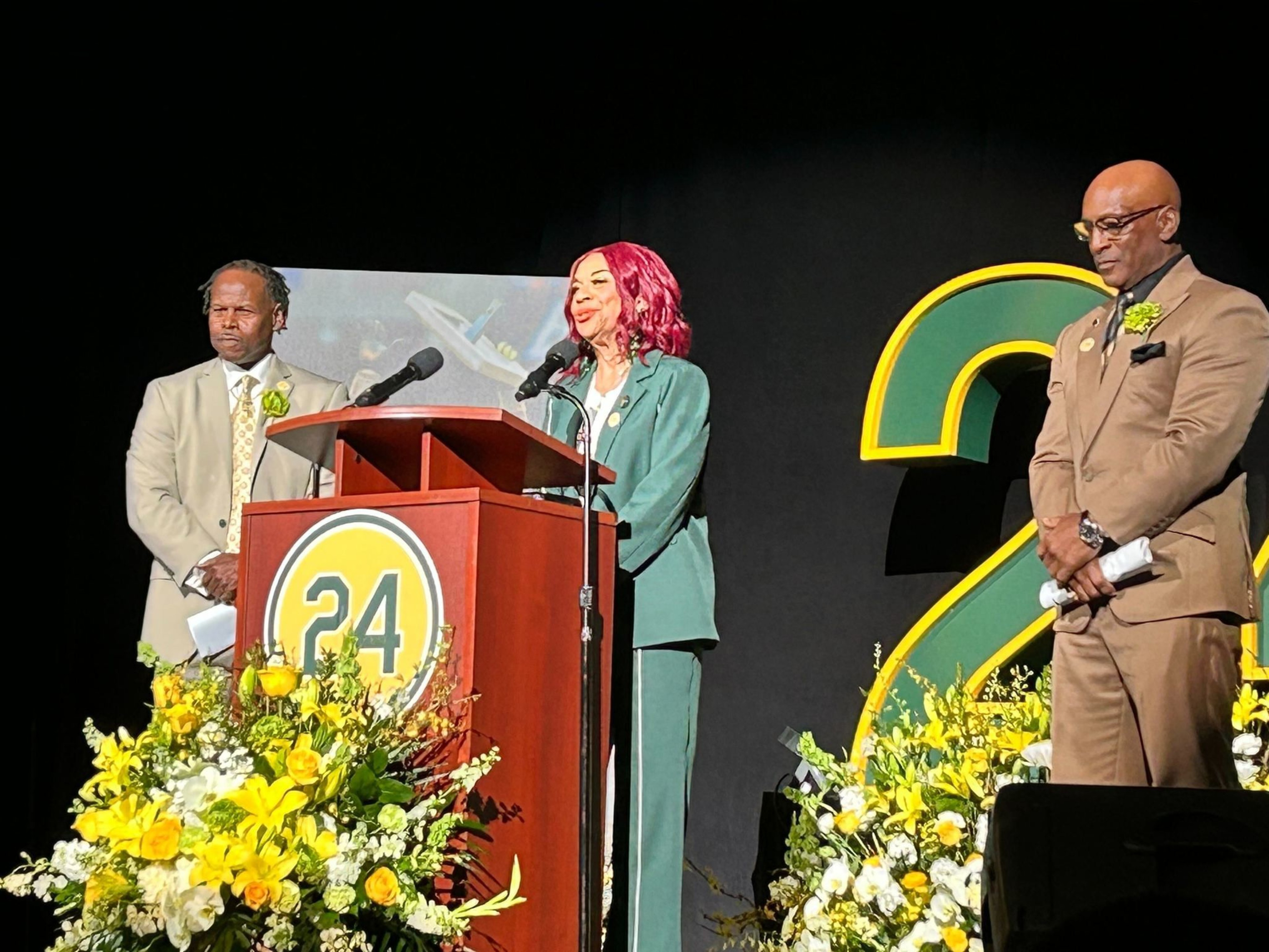 Three people stand on a stage, two men and a woman in green. They're near a podium marked &quot;24,&quot; surrounded by yellow and white floral arrangements.