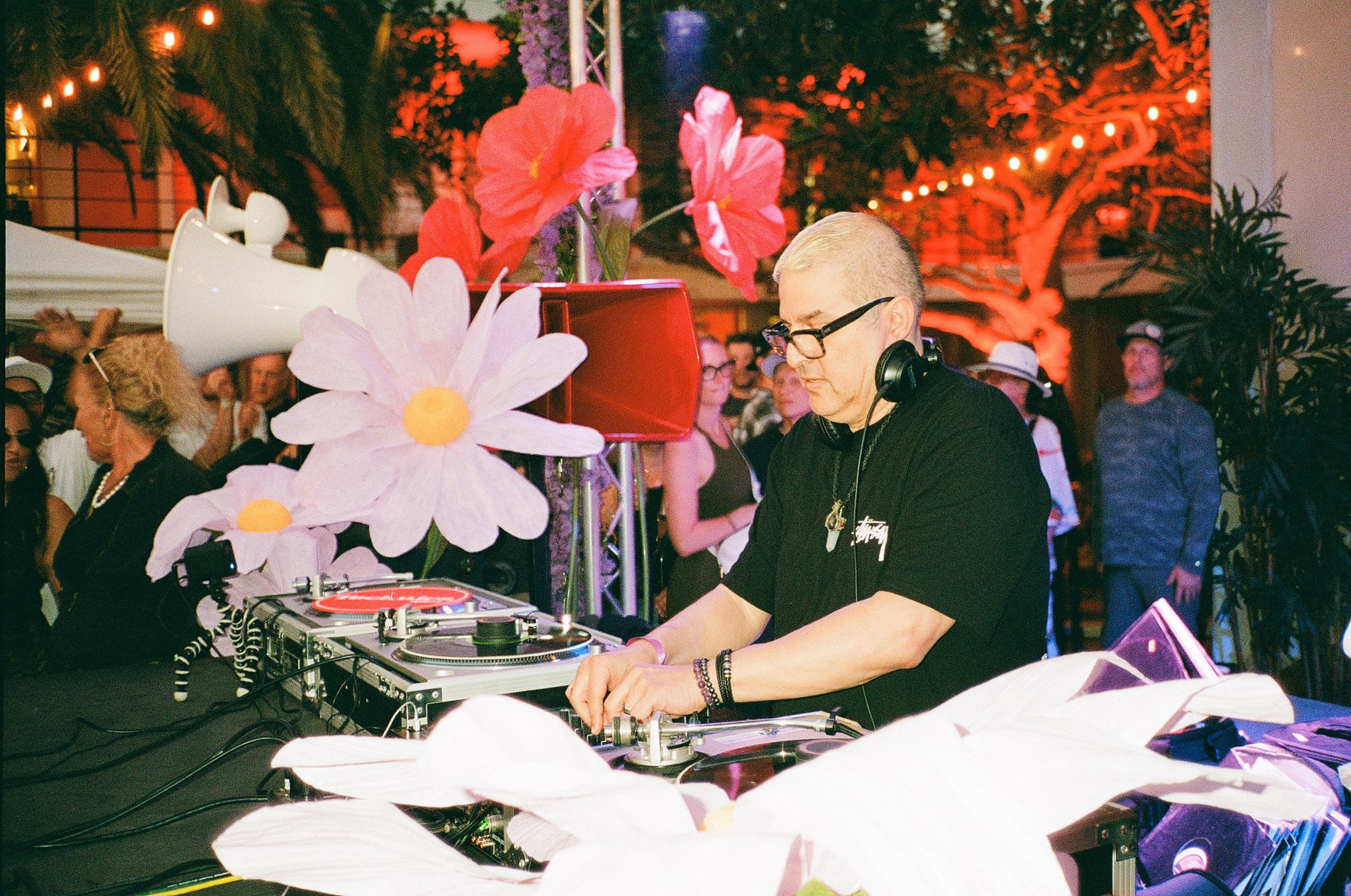 A DJ wearing glasses and headphones mixes music at an outdoor event decorated with large pink and white flower props, with a crowd in the background.