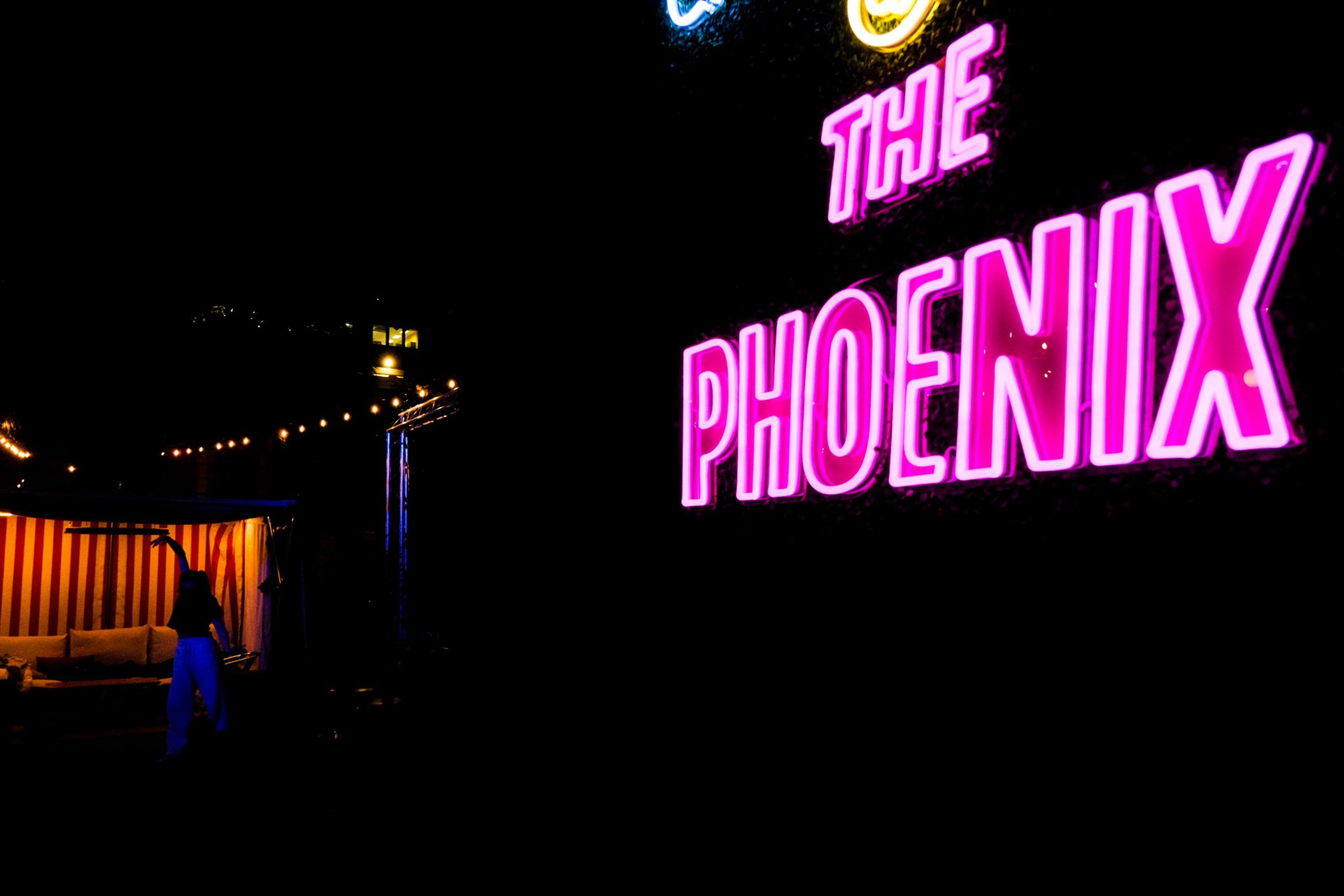 Neon sign reading "THE PHOENIX," a lit-up tent with a person, and string lights against a dark backdrop.
