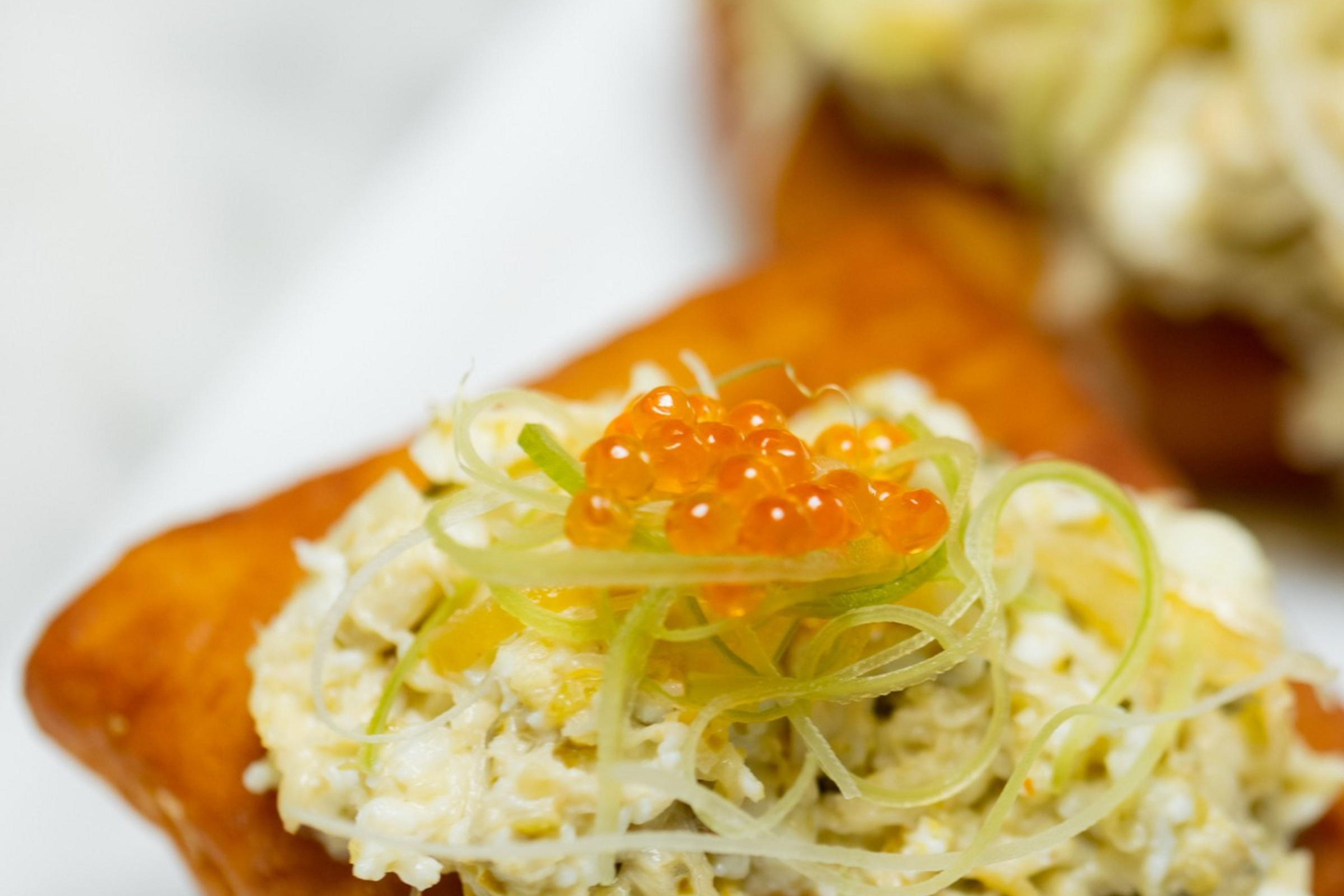 Two golden-brown pastries topped with a creamy mixture, garnished with green slivers and small orange spheres resembling fish roe, on a white plate.