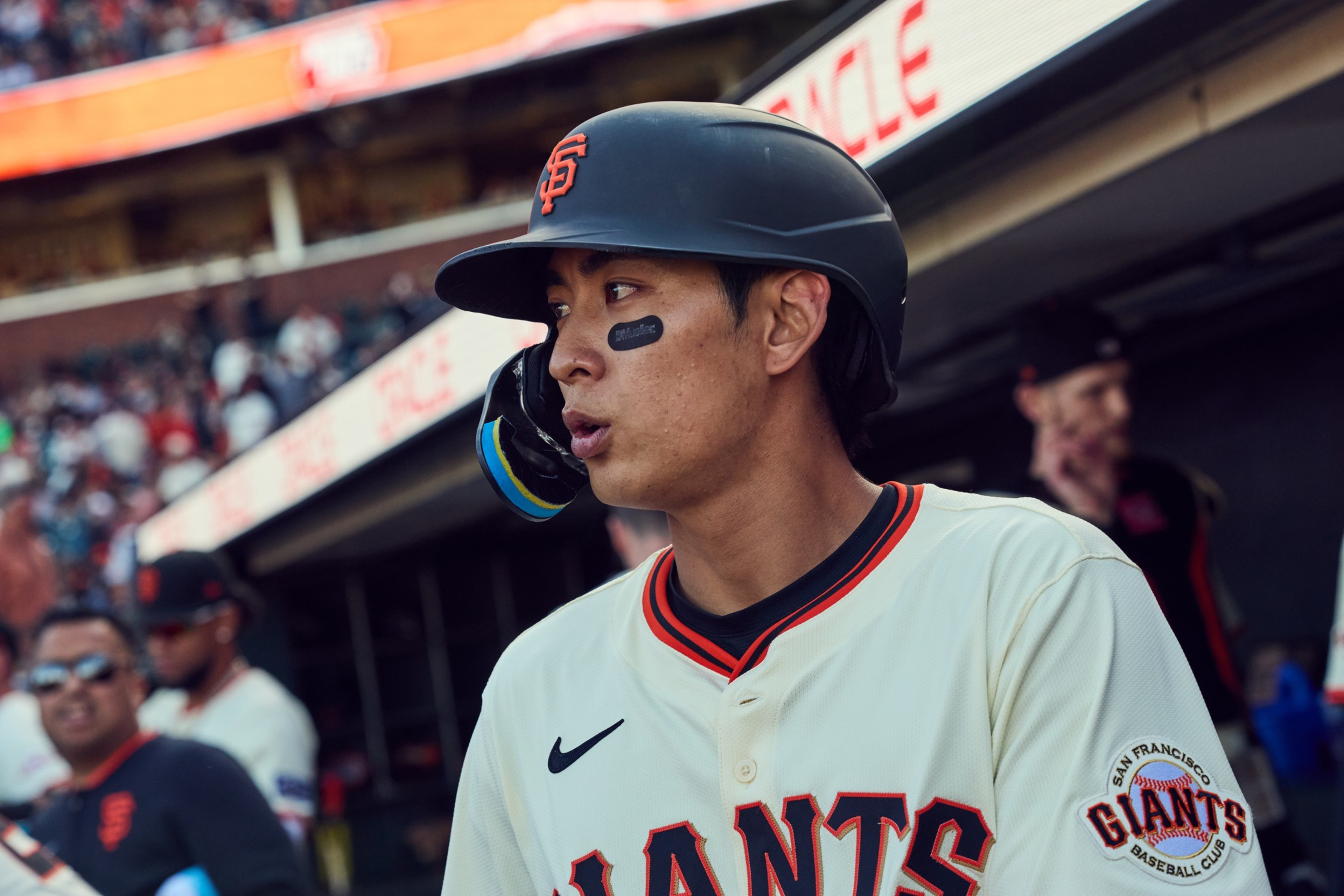 A baseball player in a Giants uniform stands near the dugout. He's wearing a helmet and eye black, with a crowded stadium in the background.
