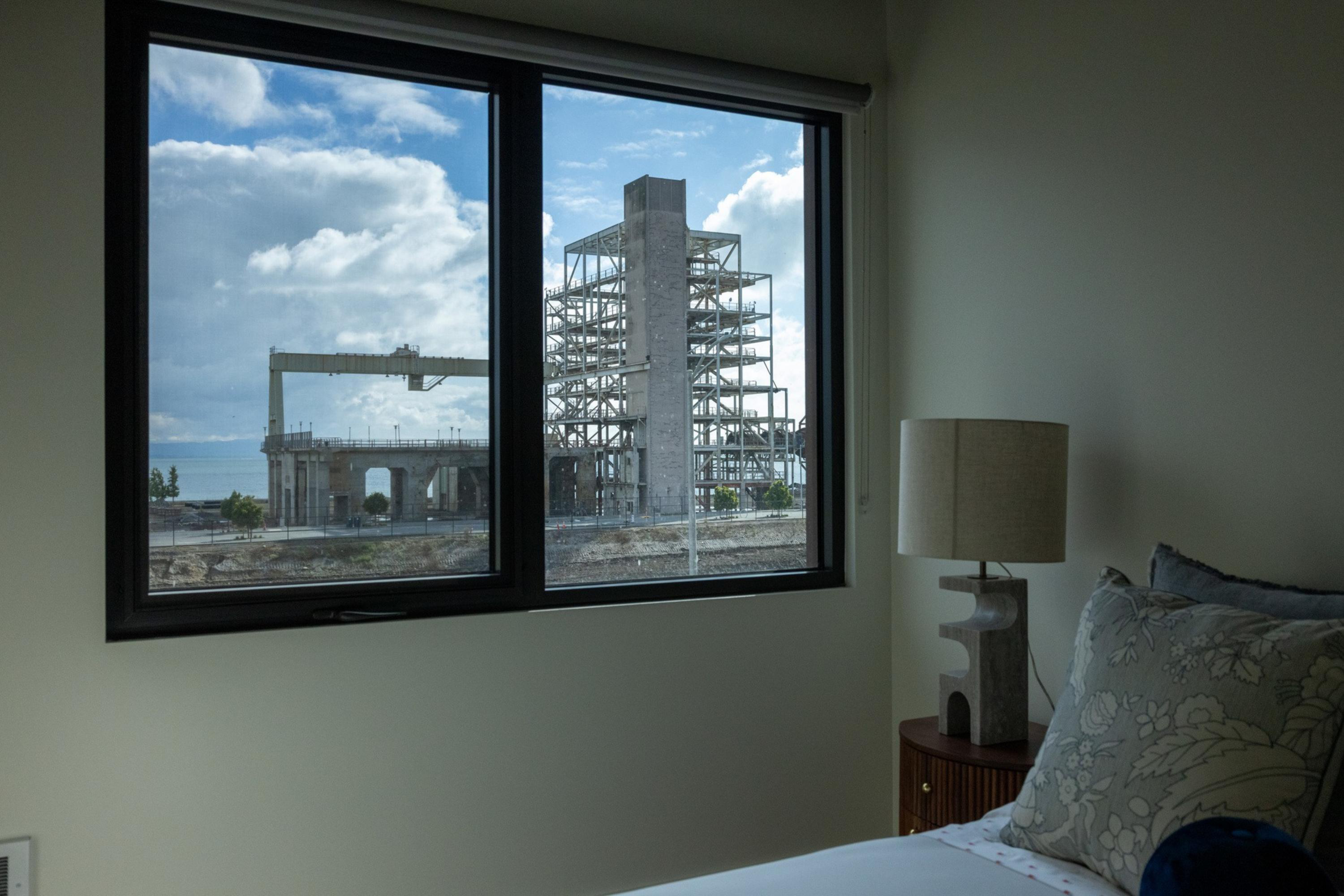 A bedroom with a bed, pillows, and a bedside table with a lamp overlooks a window showing an unfinished industrial building structure outside.