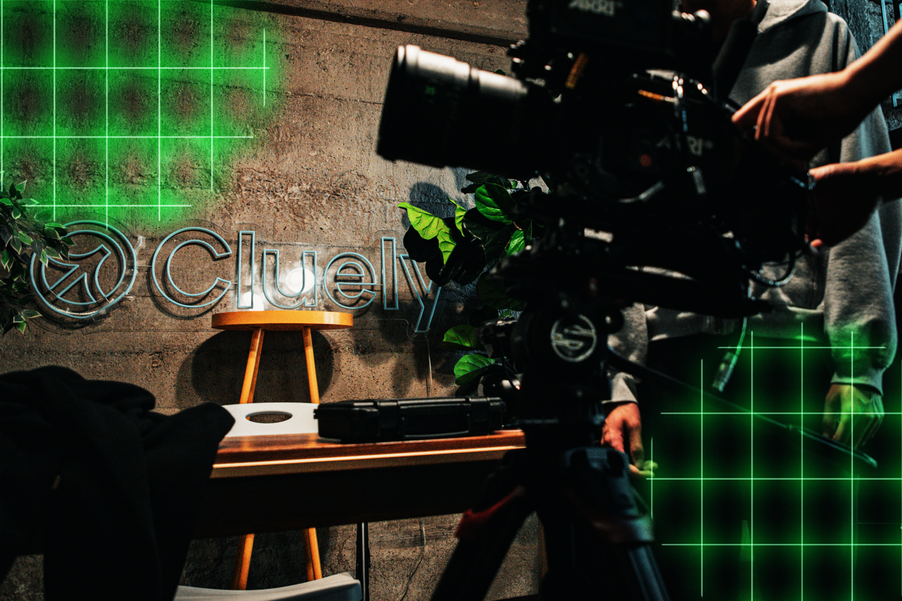 A film camera on a tripod is pointed at a wooden stool against a concrete wall with the word "Cluely" and green plants nearby.