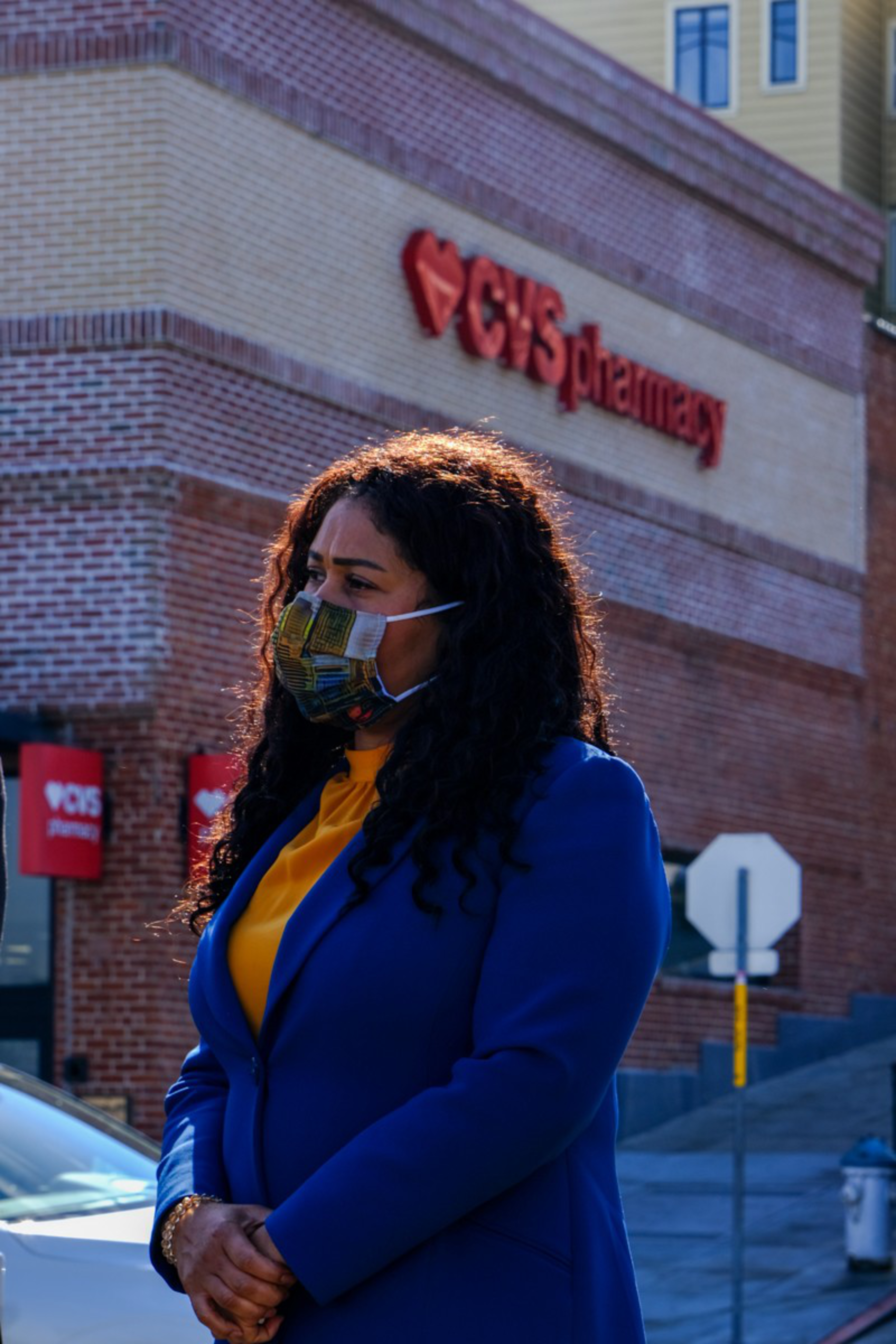 A woman wearing a patterned face mask, blue blazer, and yellow top stands outside a CVS Pharmacy building.