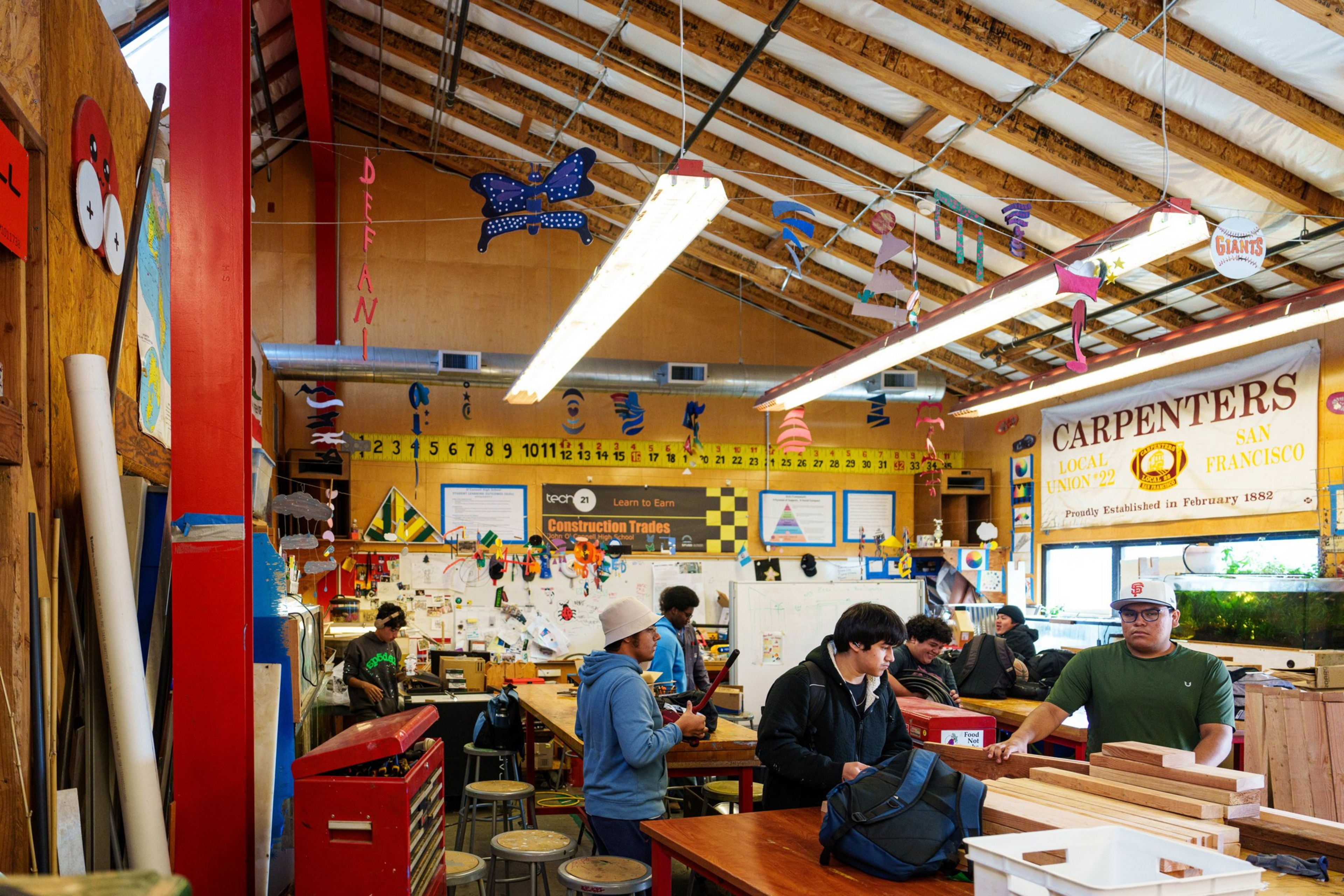 A woodworking classroom with students working, colorful crafts hanging from the ceiling, and a large &ldquo;Carpenters Local Union 22" banner on the wall.