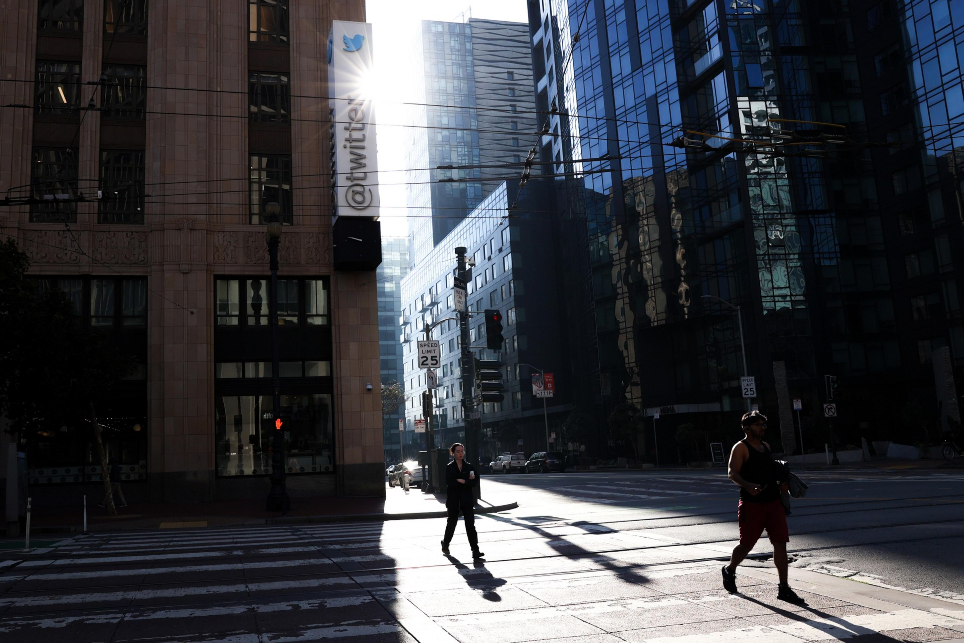 Pedestrians silhouetted against strong sunlight slanting down between tall city buildings walk alongside their own shadows as they enter crosswalks.