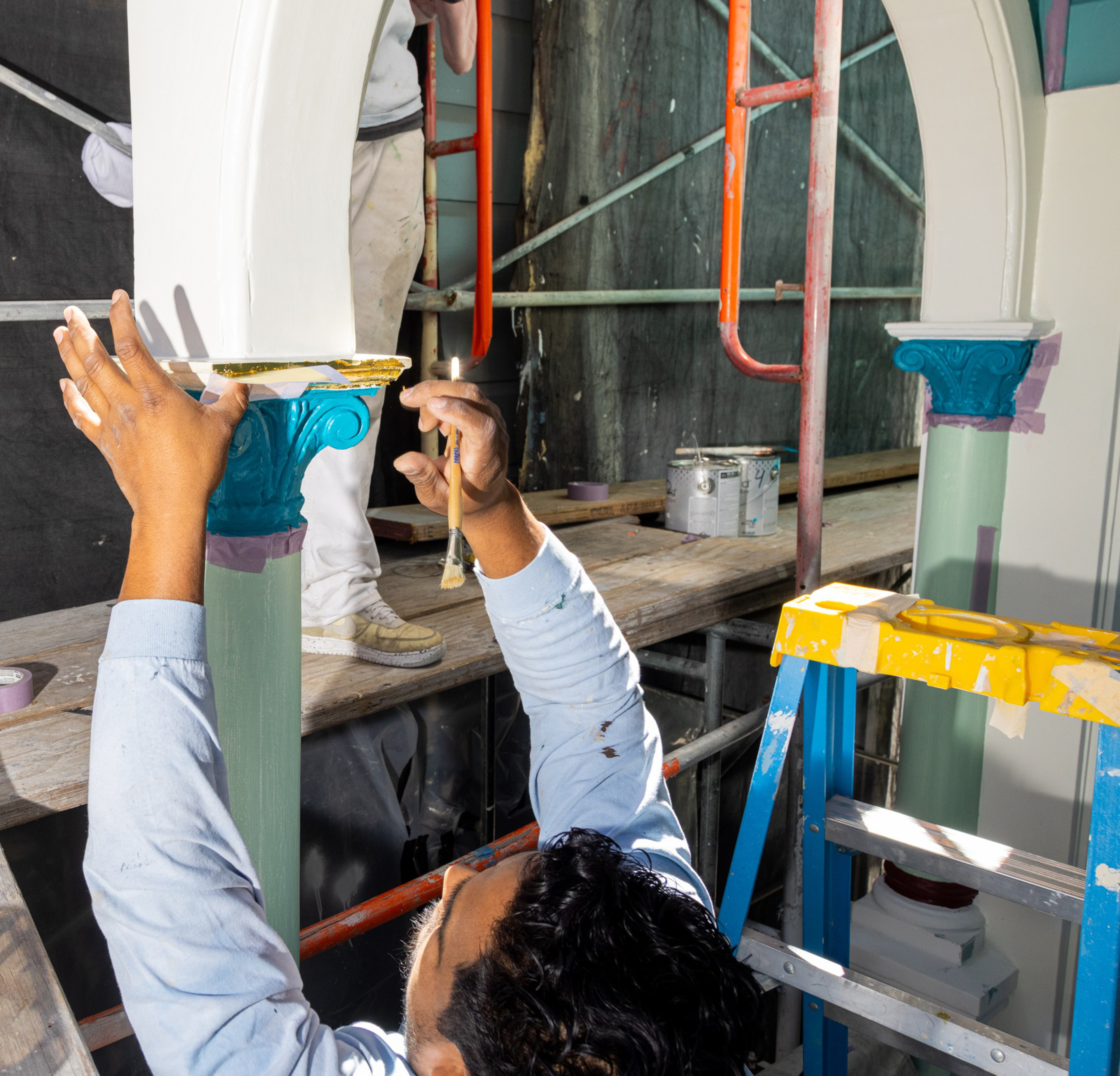 A person on a ladder painting a decorative column blue, with paint cans and scaffolding nearby.