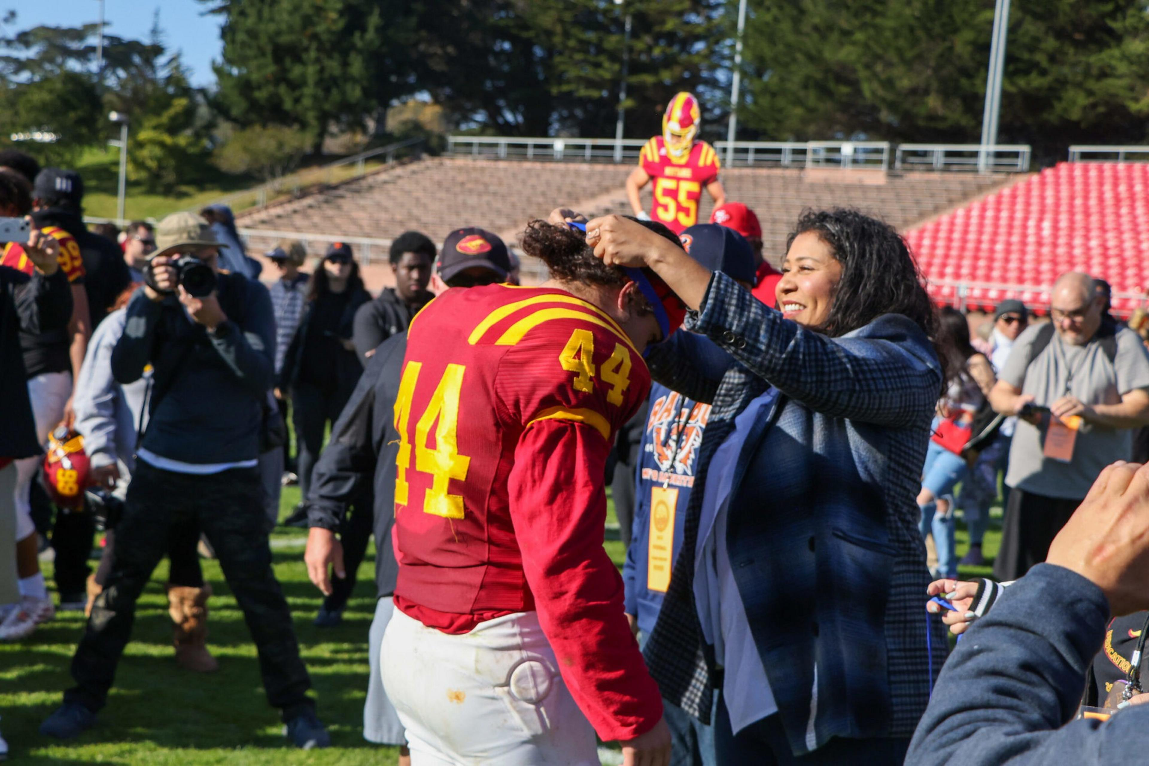 A woman places a medal on a football player wearing a red and yellow jersey numbered 44, with other players and spectators in the background.
