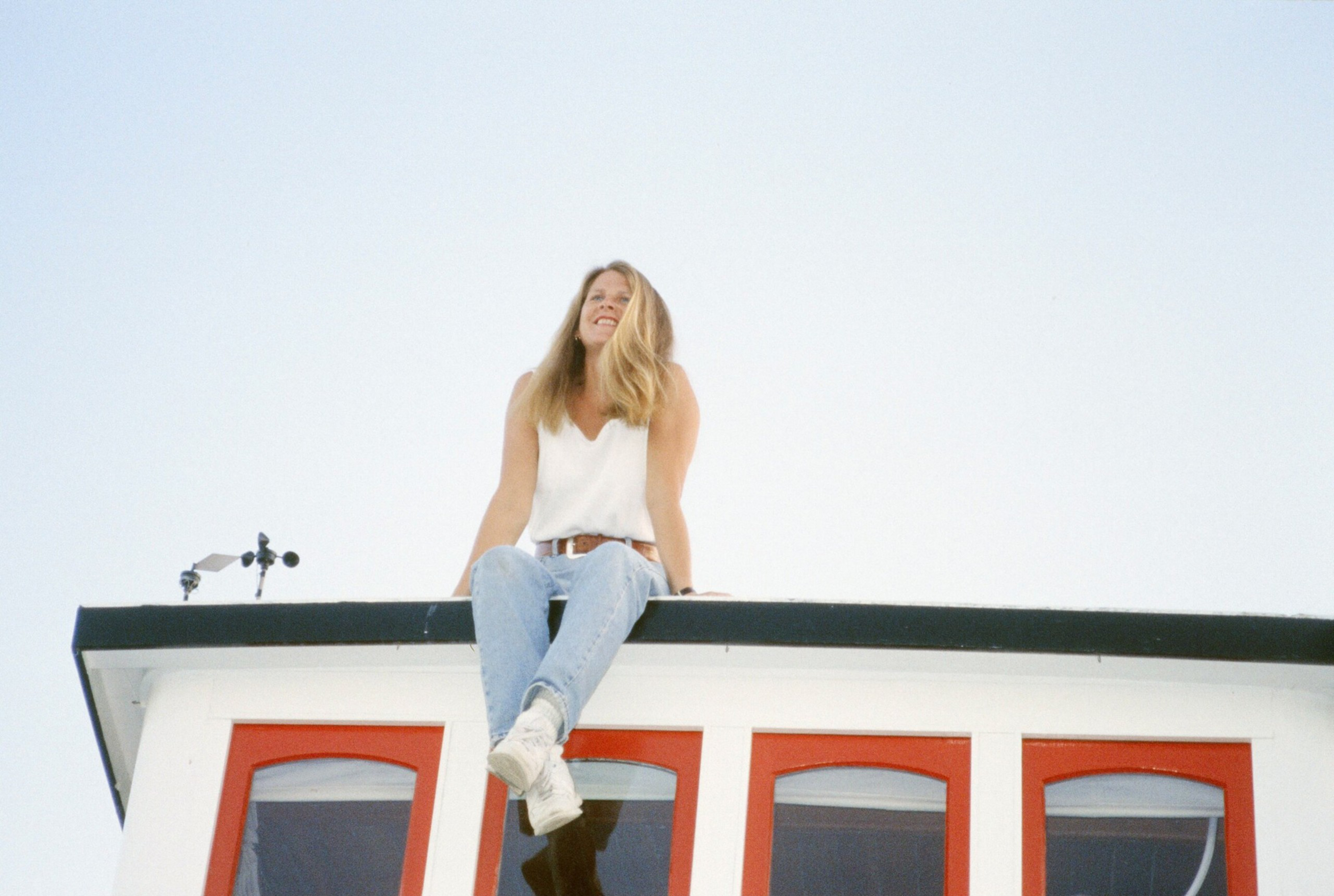 A woman with long blonde hair, wearing a white sleeveless top and light blue jeans, sits smiling on the edge of a building roof with red-framed windows.